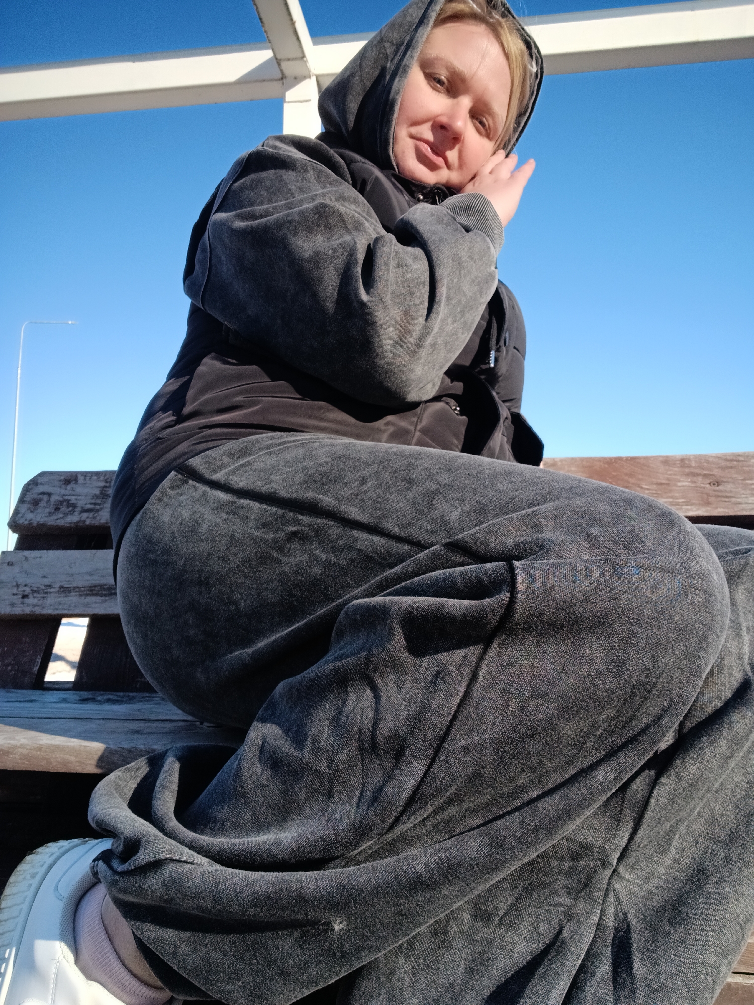 A woman with blonde hair and a grey hooded tracksuit sits on a wooden bench in Chaikovsky, Russia. She rests her left hand near her face and smiles slightly. She has her legs crossed. Her left foot, clad in a white sneaker with a grey sock peeking out, is visible at the bottom left of the frame. A streetlight is seen behind her. The sky is clear and blue. A white metal structure is visible above her head. The bench is in the background.