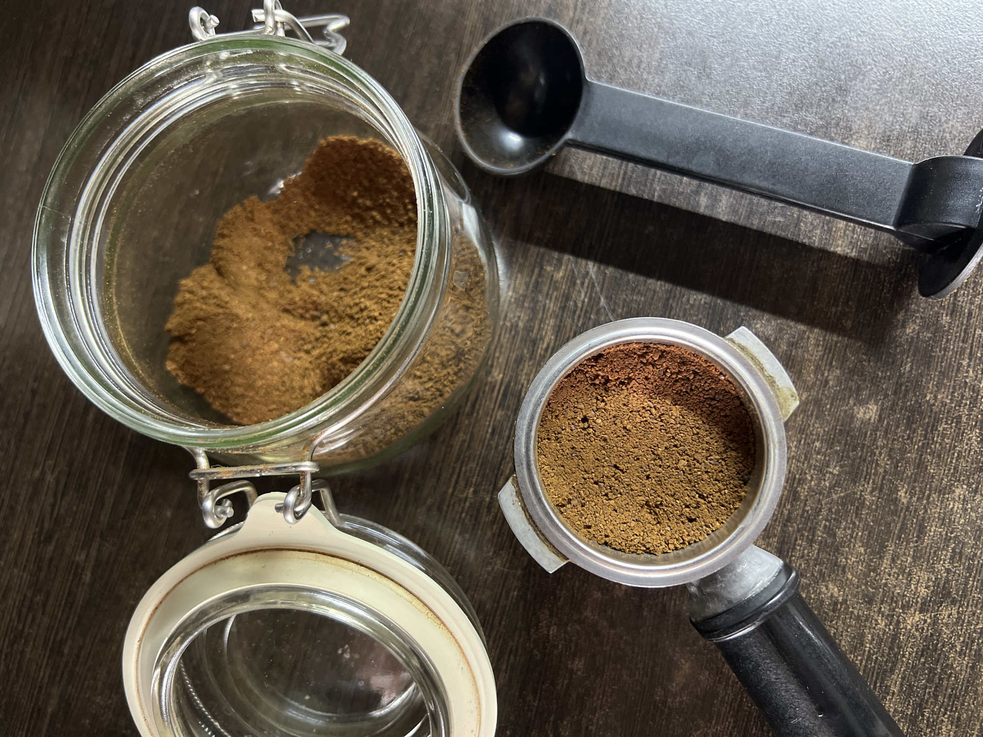 An overhead shot shows ground coffee in a clear jar with a metal clasp and white rubber seal. Beside it, a portafilter is filled with finely ground coffee. A black plastic coffee scoop lies to the right of the portafilter. The objects are placed on a dark wooden surface.