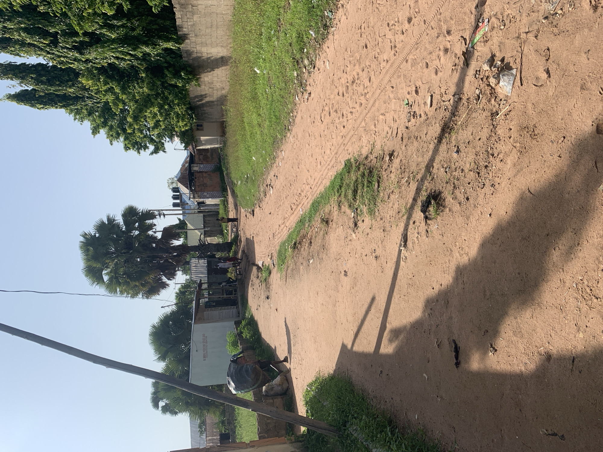 In Jalingo, Nigeria, a dirt road cuts through a residential area under a clear sky. On the left, a person carrying a large, dark bundle on their back walks away from the camera, casting a long shadow on the road. Beside them, a simple white building with text on its facade reads "M. BARKU MEDICINE STORE AND DIAGNOSTIC CENTRE". Further down the road, several people are gathered near structures with corrugated metal roofs. Palm trees and tall, slender trees line the street. On the right, a long, grey brick wall encloses a property, with lush green grass growing along its base. The road itself is sandy and worn, with tire tracks and footprints visible. Scattered debris, including plastic bags and wrappers, litters the roadside, indicating a lack of consistent waste management. The overall scene depicts a typical streetscape in a developing urban environment.