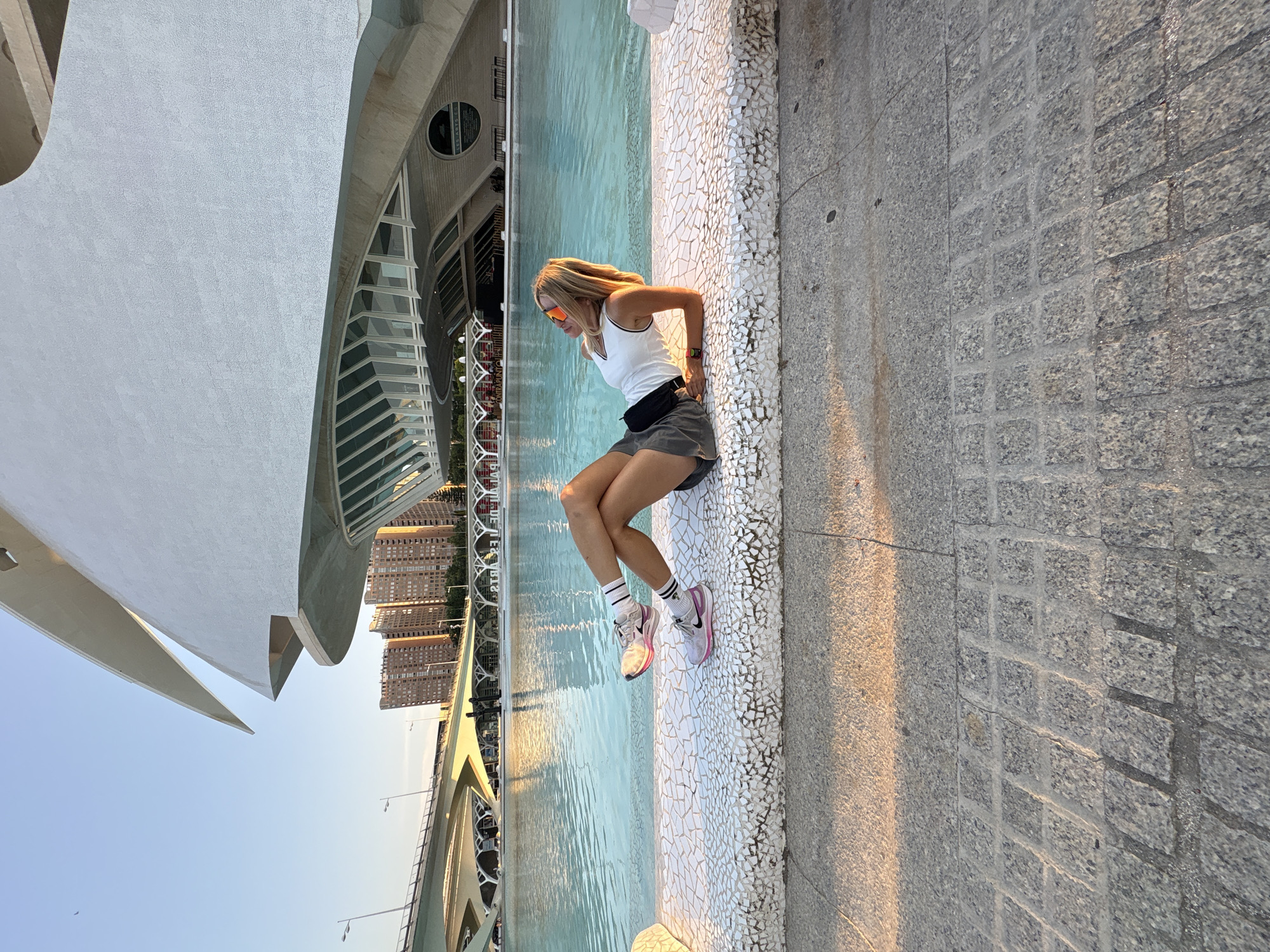 A woman with blonde hair, wearing sunglasses, a white tank top, grey shorts, and athletic shoes with pink accents, is lying on a white tiled bench. She is positioned on her left side with her legs crossed, leaning on her left elbow. The bench faces a large, calm body of blue water, reflecting the sky. Behind the water, a modern white building with a distinctive curved roofline is visible, along with a bridge and several high-rise buildings. The scene appears to be set outdoors during the day, with the sun casting shadows and warm light on the ground. This location is the City of Arts and Sciences in Valencia, Spain, specifically the area in front of L'Hemisfèric.