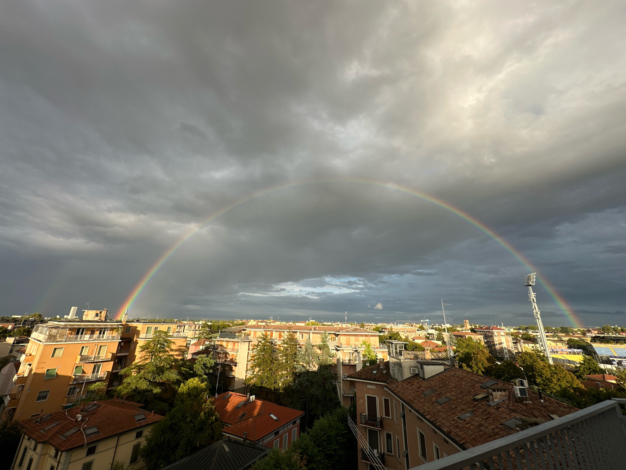 This is an aerial view of a city with buildings and trees under a dramatic sky. A rainbow is prominently displayed, arching across the frame. The sky is a mix of dark, stormy clouds and areas of bright sunlight, creating a stark contrast. Buildings with tiled roofs and balconies are visible, some with trees interspersed between them. A tall stadium light structure is seen on the right side of the image. The scene appears to be taken from a high vantage point, likely a balcony or rooftop, looking out over a residential and possibly urban area. The overall impression is one of a city experiencing a weather transition, with the beauty of a rainbow emerging after or during a rain shower. The architecture suggests an Italian setting, consistent with Parma.
