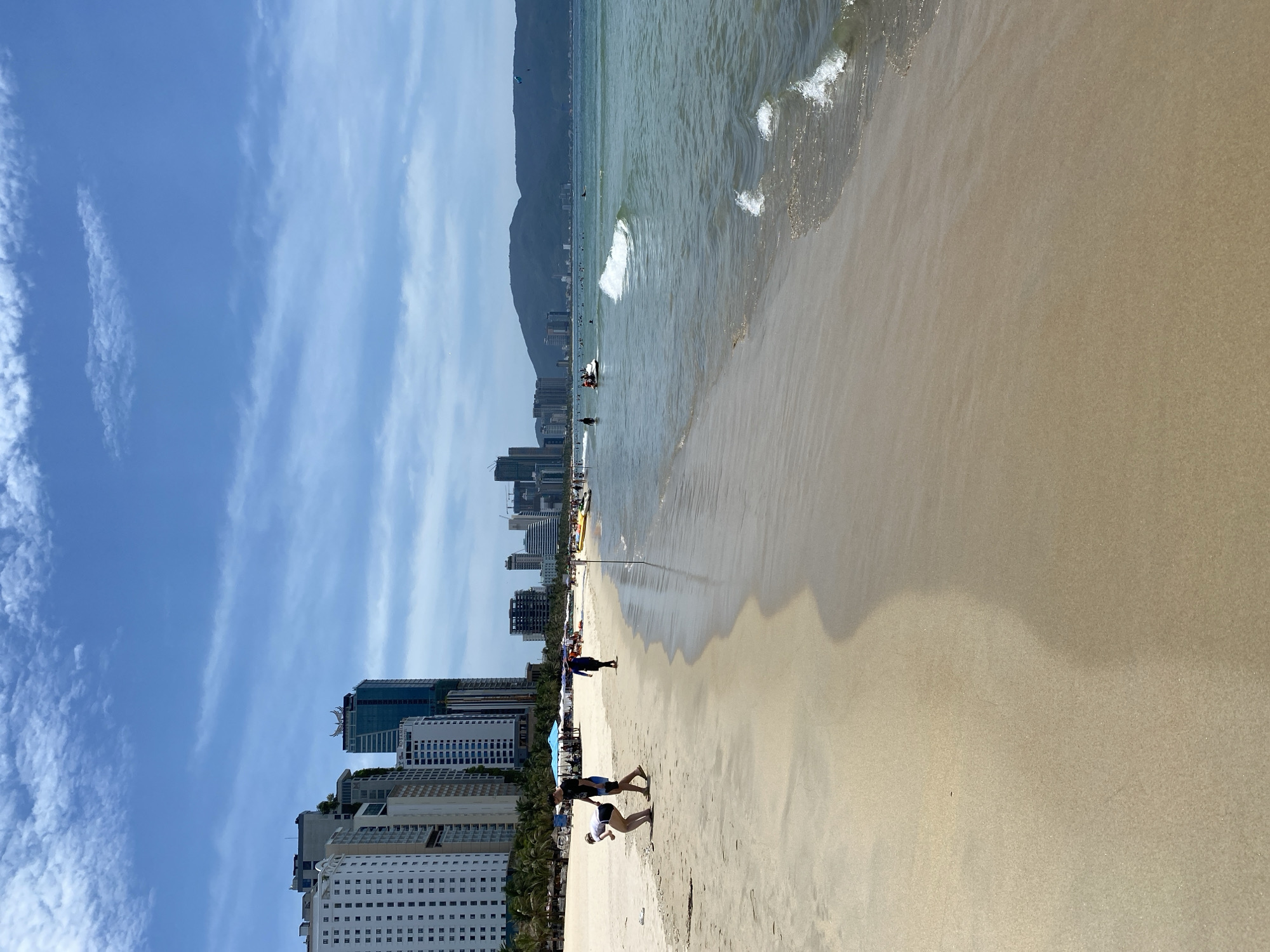 This is a vertical, eye-level, medium shot of a beach in Đà Nẵng, Vietnam, on a bright, sunny day with a partly cloudy sky. The foreground is dominated by a wide expanse of light-colored sand, wet near the water's edge. Gentle waves with white foam lap at the shore.
In the mid-ground, two people are visible on the sand. A woman in a white top and black shorts is bending down, appearing to pick something up from the sand, while a man in a black t-shirt and blue shorts stands nearby, looking in her direction. Further down the beach, a person in a dark outfit walks away from the viewer. A few beach umbrellas are set up further back on the sand.
The background features the skyline of Đà Nẵng. Several tall, modern buildings, including high-rise hotels and apartments, line the coast. Beyond the city, a range of green, hilly mountains forms a backdrop. The ocean is a clear, greenish-blue color. Several boats and jet skis are visible in the water, and a few people are in the ocean. A paraglider is visible in the far distance. The overall impression is a lively and pleasant day at the beach.