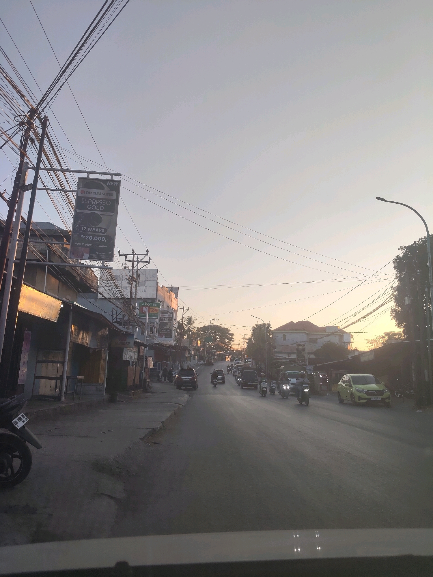 The image depicts a street scene in Kupang, Indonesia, likely during the late afternoon or early evening, judging by the low angle of the sun and the soft, warm light. The street is lined with buildings, some appearing to be shops or small businesses, with signage visible in Indonesian. Power lines are a prominent feature, crisscrossing the sky and supported by utility poles. A variety of vehicles, including cars and numerous motorcycles, are present on the road, suggesting moderate traffic. The road surface appears somewhat worn in places. The sky is a pale, hazy blue, with a gradient towards a warmer hue near the horizon, indicating the sun is either rising or setting. One building has a sign for "DJARUM SUPER ESPRESSO GOLD" advertising 12 wraps for Rp 20,000,-. Another sign indicates "APOTEK 24," suggesting a 24-hour pharmacy. A green car is parked on the right side of the street, and several motorcycles are in motion. The overall impression is of an active urban environment with functional, albeit somewhat aged, infrastructure.