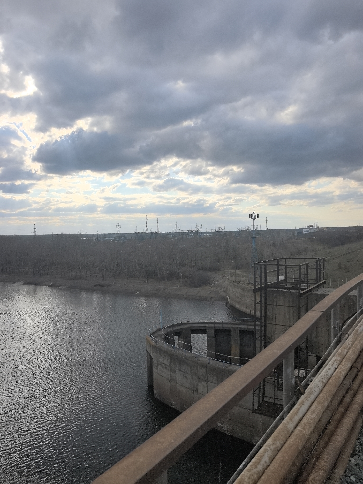 The image captures an expansive, somewhat stark industrial landscape featuring a large body of water under an overcast sky. In the immediate foreground on the right, a rusty metal railing runs diagonally, with a pile of weathered, corroded pipes visible beneath it.
The main subject in the midground is a large, curved concrete structure extending into the dark water, reminiscent of a dam's spillway or a water intake tower. This structure features a metal platform and a tall, blue-painted mast with lights or communication equipment. The water surface is dark and shows subtle ripples, indicating movement.
The far bank of the water is lined with a dense row of bare, deciduous trees, suggesting the season is late autumn, winter, or early spring. Beyond the treeline, the flat horizon is dominated by a sprawling industrial complex. Numerous high-voltage electricity pylons are visible across the distant landscape, connecting to large industrial buildings or power plants.
The sky is heavily overcast with thick, grey clouds, although brighter patches and glimpses of sunlight are visible towards the left, suggesting a break in the cloud cover. The overall impression is one of a functional, utilitarian environment, possibly a power generation or water management facility.
No people are visible in the scene, and there is no discernible text. The image depicts a cold, industrial day in Chaikovsky, Russia.