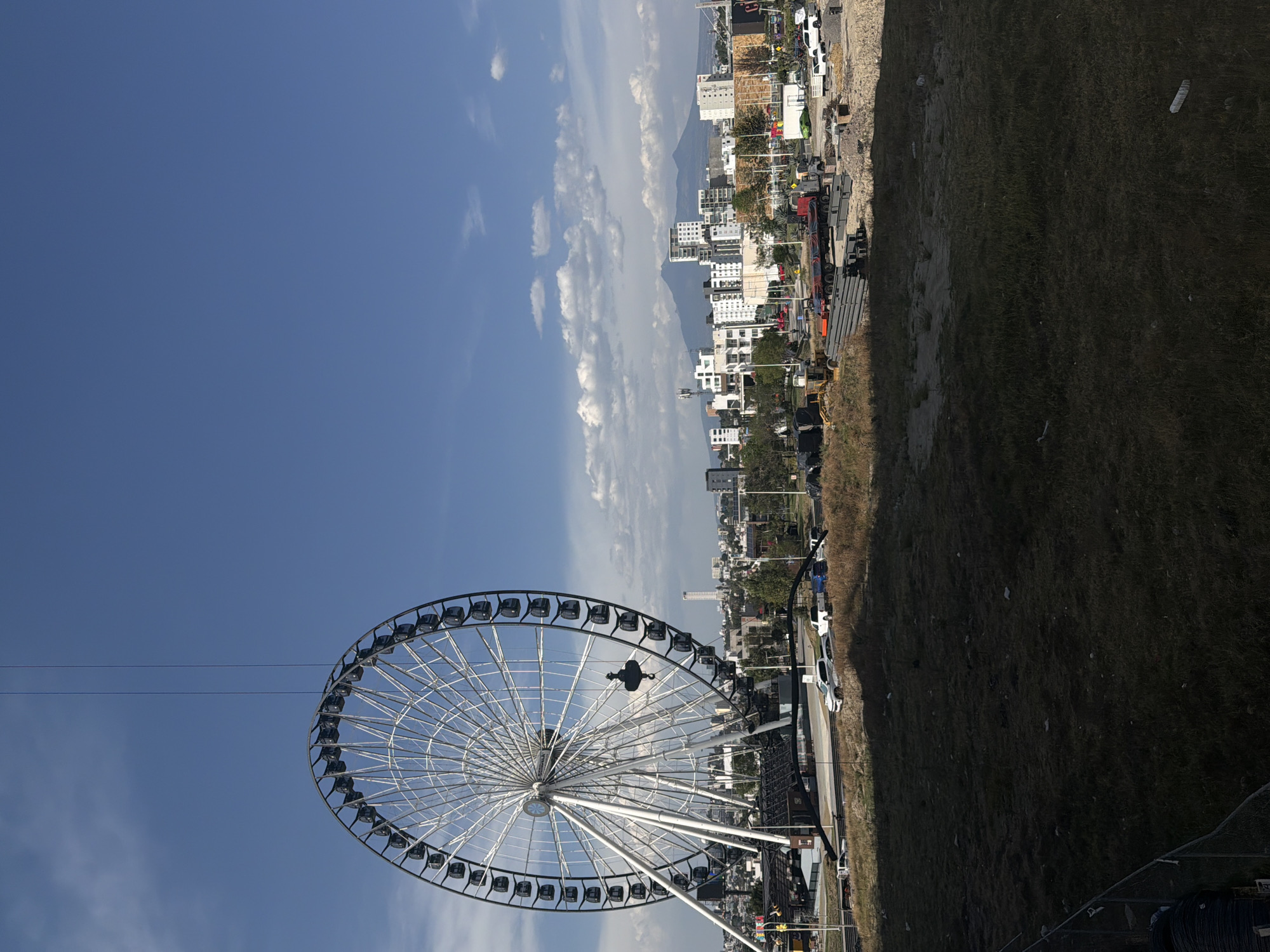 The image captures a Ferris wheel in front of a city skyline under a partly cloudy sky. The Ferris wheel is prominently featured, with its structure and passenger cars visible. Behind it is a cityscape with buildings of varying heights and styles. In the background, a mountain can be observed. A road with vehicles is also visible. The foreground consists of what appears to be grass and some construction or industrial materials.