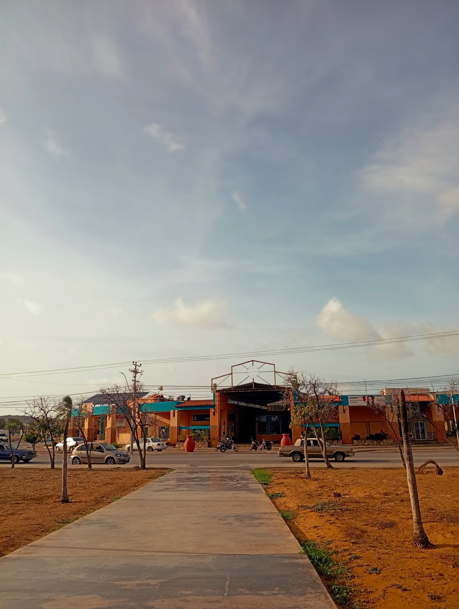 The image captures a street scene in Juan Griego, Venezuela, featuring a long, concrete walkway in the foreground. To the right of the walkway is a patch of dry, reddish-brown soil with sparse green grass and a few trees. Beyond the soil, a road is visible with several vehicles, including cars and motorcycles, driving or parked.
In the background, a commercial building complex with an orange and blue facade lines the street. The building has an open-air covered section with a metal structure. Trees with bare branches are planted in front of the building, and a large orange ceramic pot is placed near the entrance. Power lines stretch across the sky above the buildings. The sky is a light blue with scattered white clouds. The overall impression is a sunny day in a coastal town, with a mix of natural and urban elements.