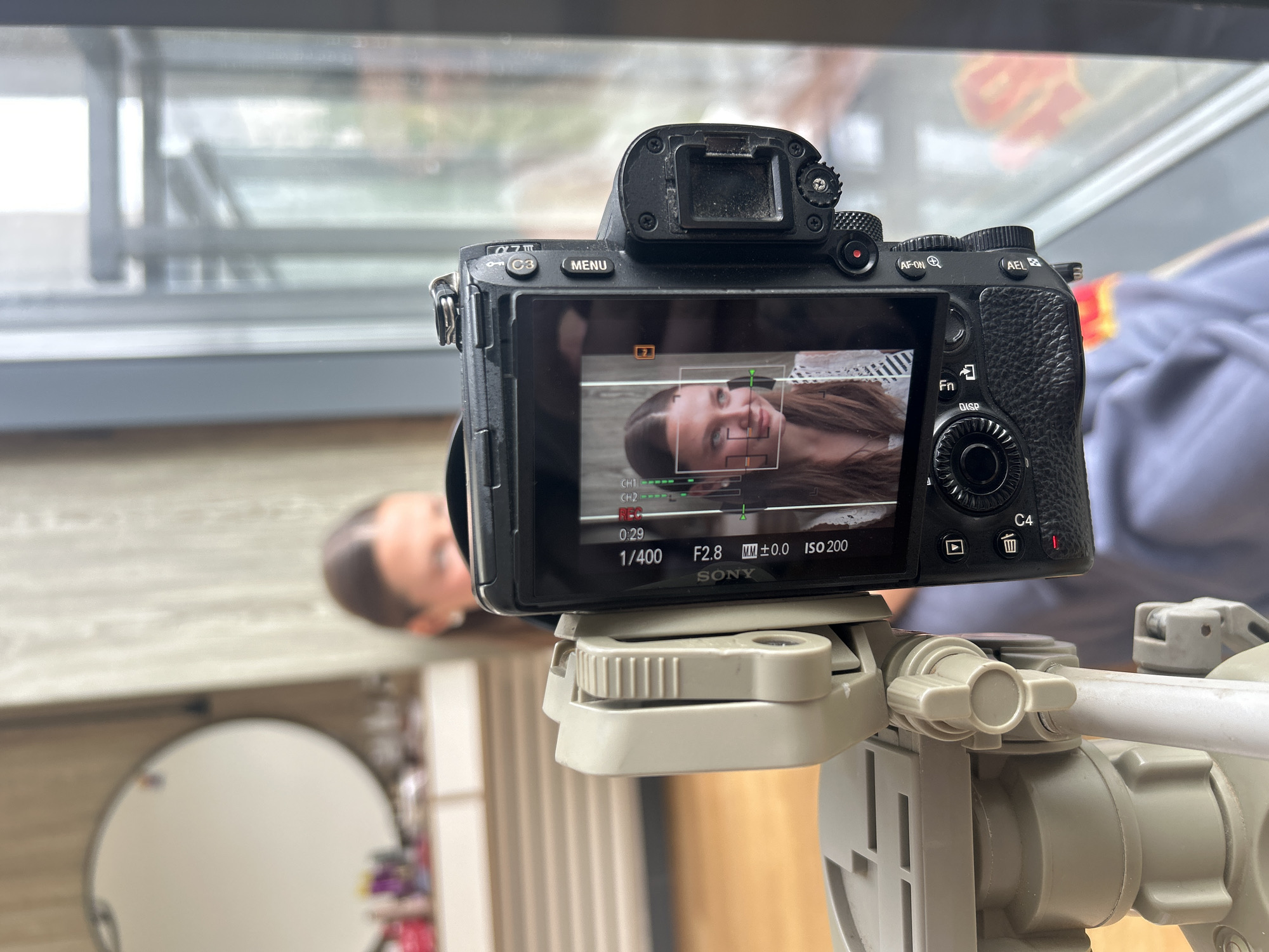 A black Sony camera, likely an Alpha series (based on the "α7 III" marking), is mounted on a grey tripod. The camera's LCD screen displays a portrait of a young woman. The camera settings displayed on the screen show a shutter speed of 1/400, an aperture of f/2.8, and an ISO of 200. In the background, there's a blurry view of a window frame and the vague reflections outside. Part of a mirror is also visible. The location cannot be discerned from the image.