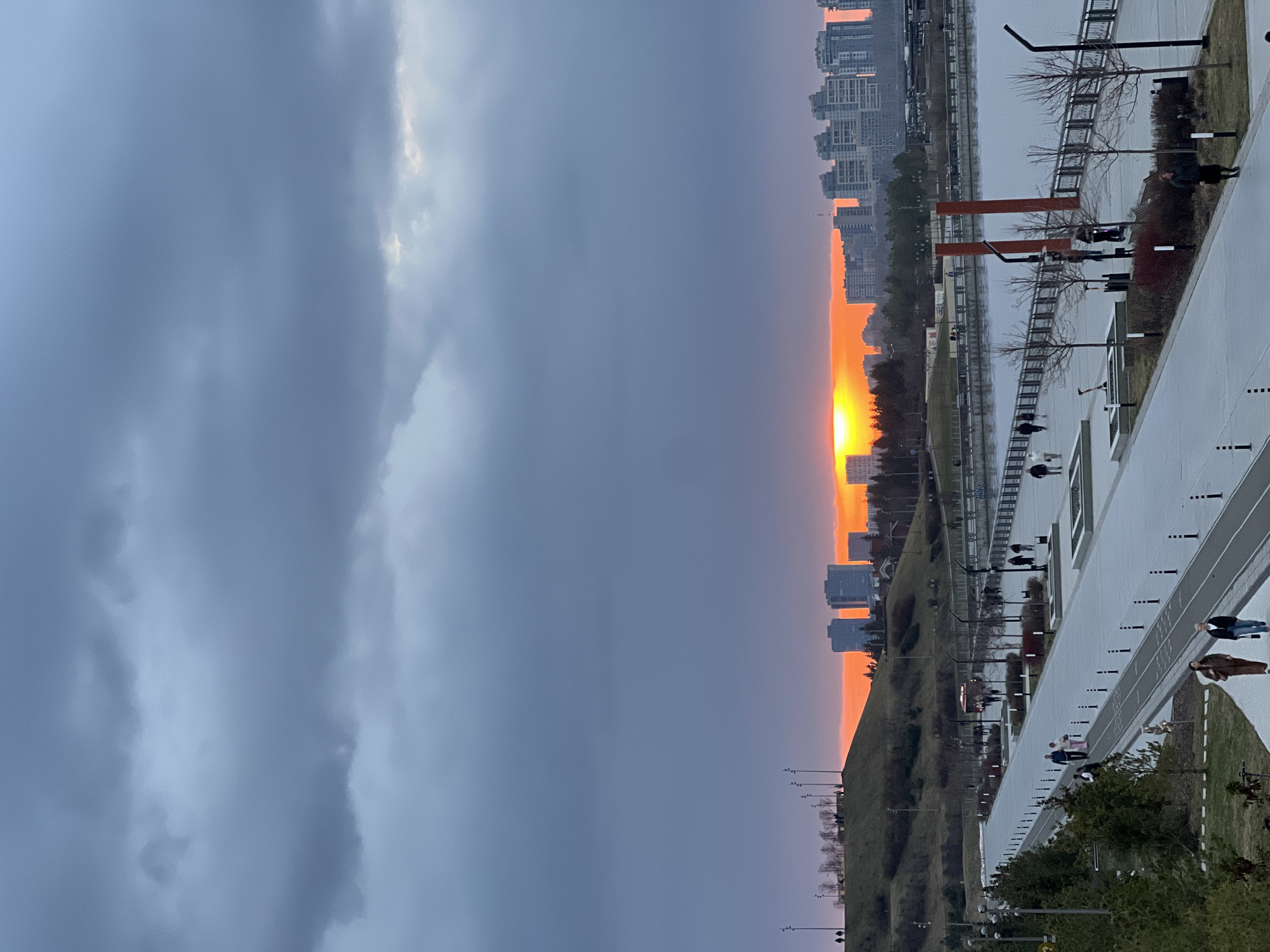 The image shows a sunset in Kazan, Russia. The sky is predominantly overcast with dark, ominous clouds covering most of the frame. A vibrant orange sunset is visible on the horizon, framed by a thin band of buildings.
A long, white walkway stretches across the bottom right of the frame. People are walking along the walkway. A grassy slope descends from the walkway. Modern buildings are visible across the water. The picture is taken at eye level.