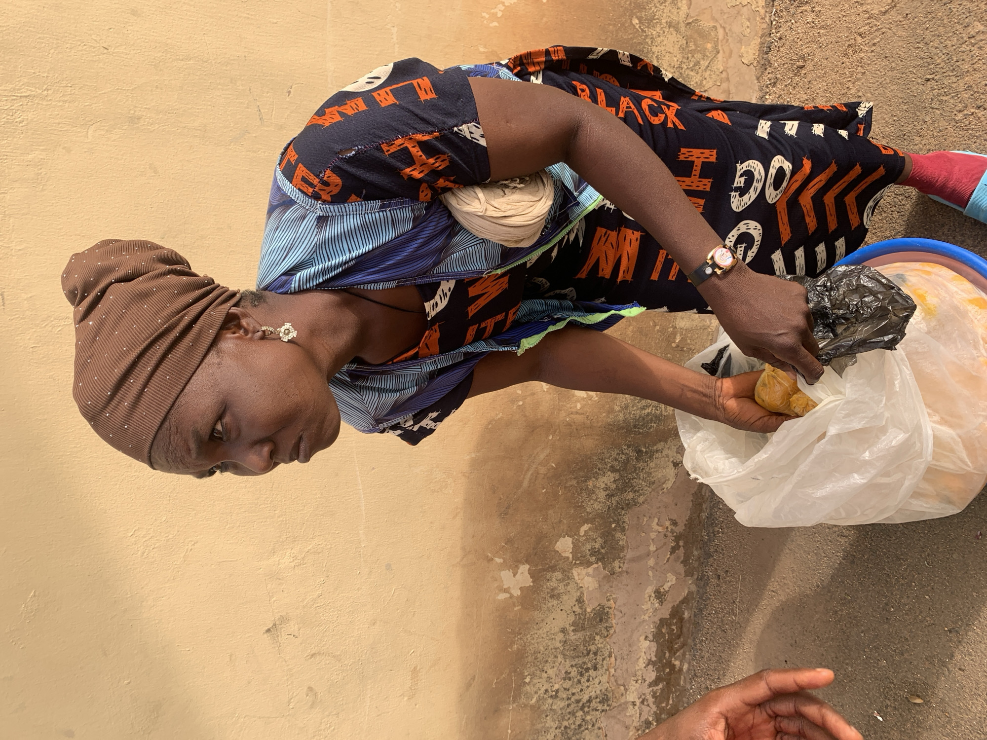 An adult female wearing a brown headscarf, a dress with blue striped and black patterned elements, and a watch is situated outdoors in what is likely the city of Bukuru, Nigeria, given the subject's complexion and attire. She is holding a white plastic bag that contains yellow items. Her left hand reaches into a black plastic bag. The background includes a yellow wall and worn ground.