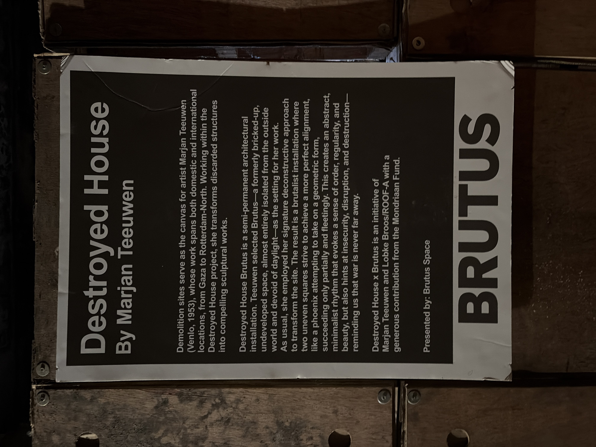 The image shows a poster with the title "Destroyed House" by Marjan Teeuwen. The poster is displayed on a wooden surface, possibly a wall or a panel. The poster is dark with white text. The text describes the artist's work, which involves transforming discarded structures from demolition sites into sculptural works. It specifically mentions the "Destroyed House Brutus" installation, which is a semi-permanent architectural installation in an undeveloped space. The poster also states that "Destroyed House x Brutus" is an initiative of Marjan Teeuwen and Lobke Broos/ROOF-A with a generous contribution from the Mondriaan Fund. It is presented by Brutus Space. At the bottom of the poster, the word "BRUTUS" is displayed in large, bold letters. The overall scene appears to be an indoor exhibition space, with the poster serving as an informational display for an art installation.