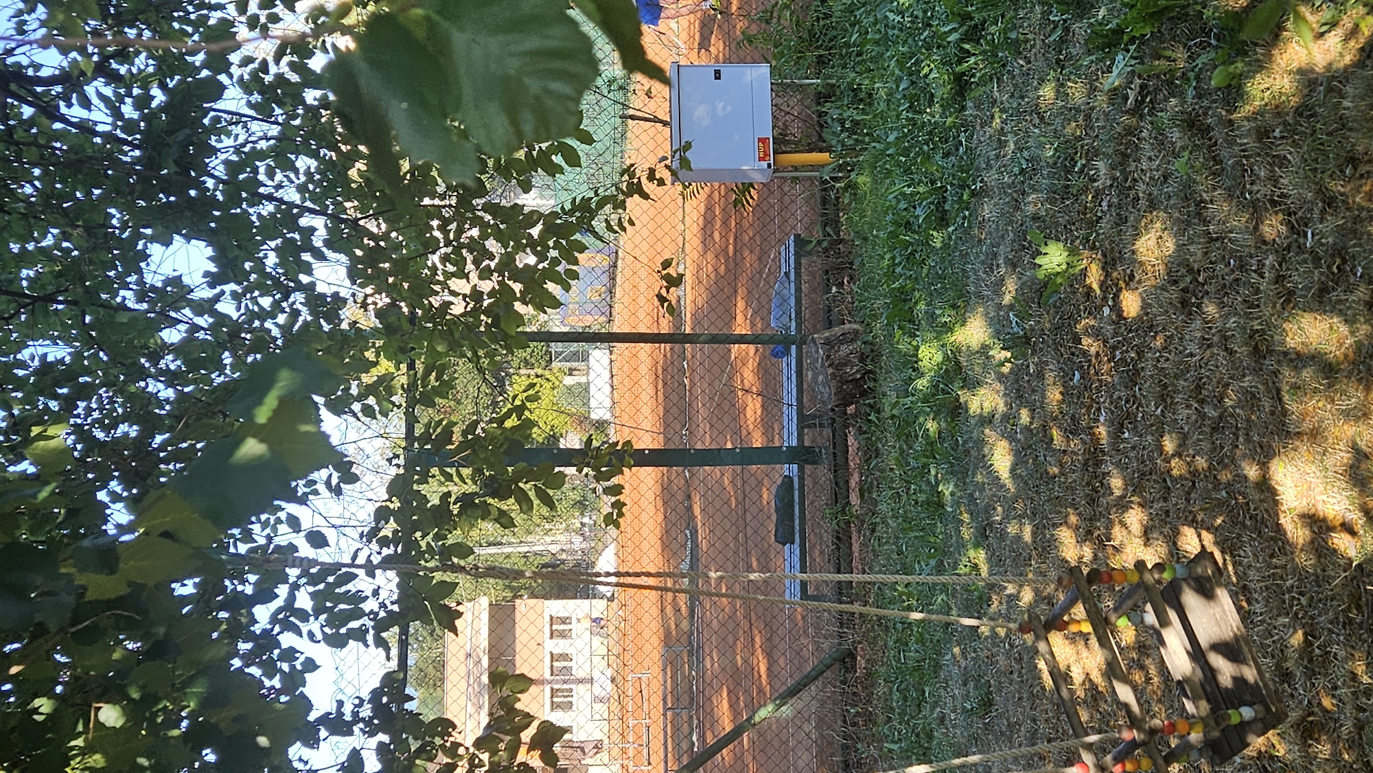 The image depicts a view from under a leafy tree canopy, looking out onto a tennis court in Prešov, Slovakia. In the foreground, a wooden swing set with colorful beads on its ropes is visible. The ground beneath the swing is dry grass with dappled sunlight. Beyond the swing, a chain-link fence encloses a clay tennis court. A green metal bench with two dark objects on it is situated near the fence. In the background, a building with multiple windows is visible, and further back, more of the tennis court and trees can be seen. A white electrical box with a yellow pole is mounted near the fence.