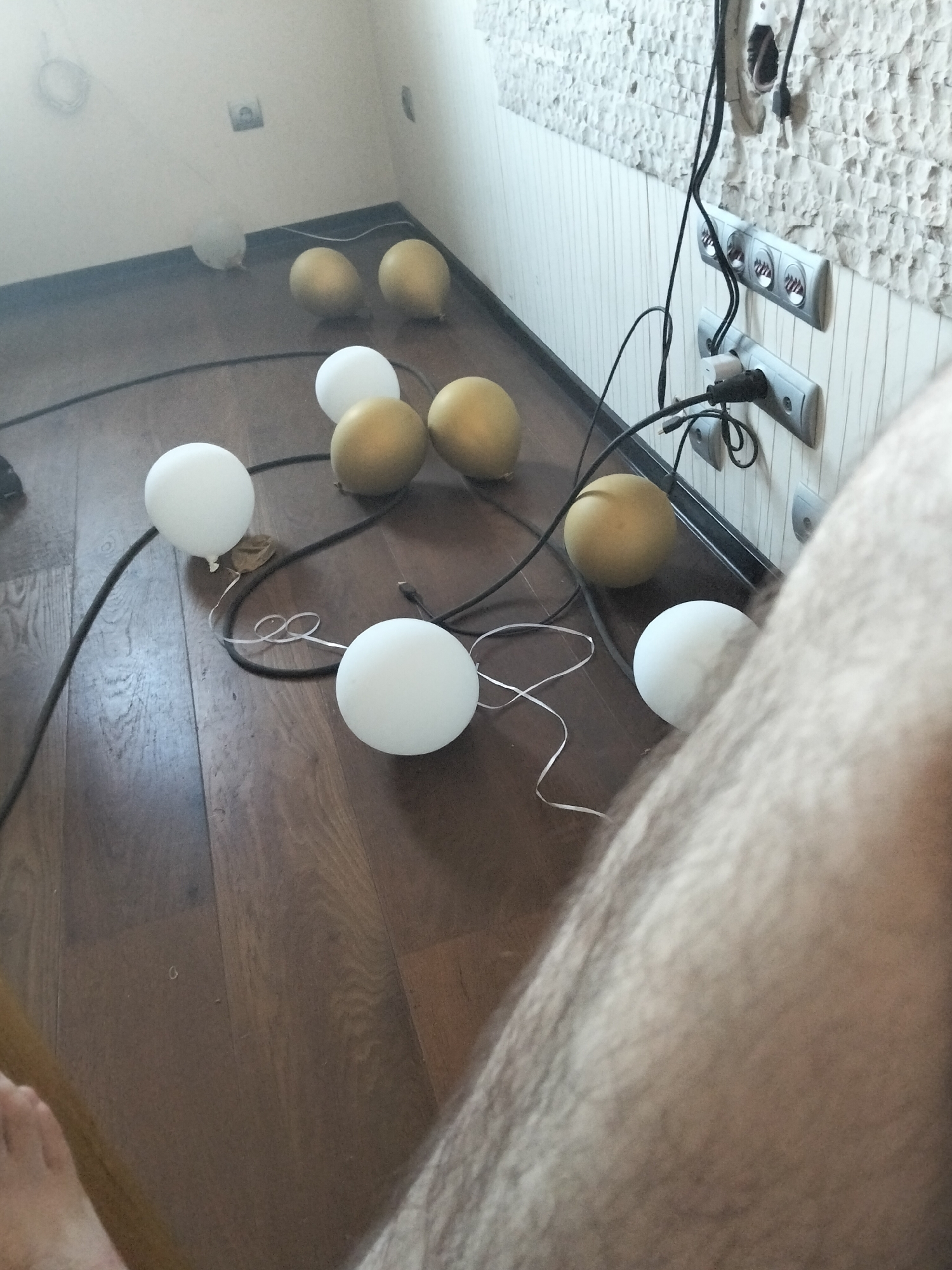 The image displays a floor with several balloons scattered across it, some white and some golden. A black cord runs across the floor, connecting to some of the balloons. In the background, there are electrical outlets and wires on a textured wall. A portion of a person's leg and foot is visible in the foreground, suggesting they are either sitting or lying down. The overall environment appears to be an indoor space, possibly a room in a home or apartment.