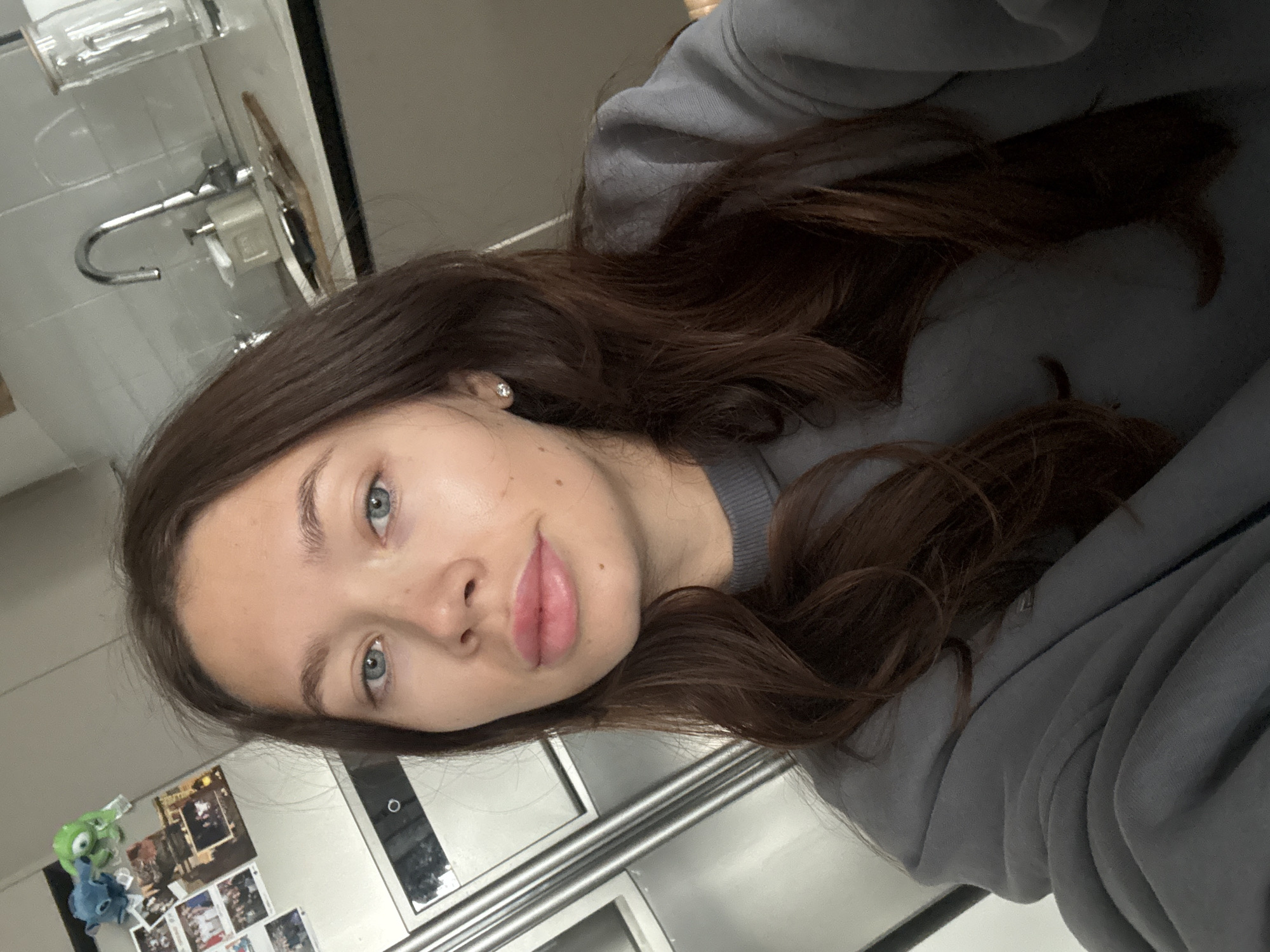 A woman with brown hair and blue eyes is posing in a kitchen. She is wearing a gray sweater. A faucet is visible in the background. A refrigerator with magnets and photos on it is also visible. The location cannot be determined as Tel-Aviv, Israel.