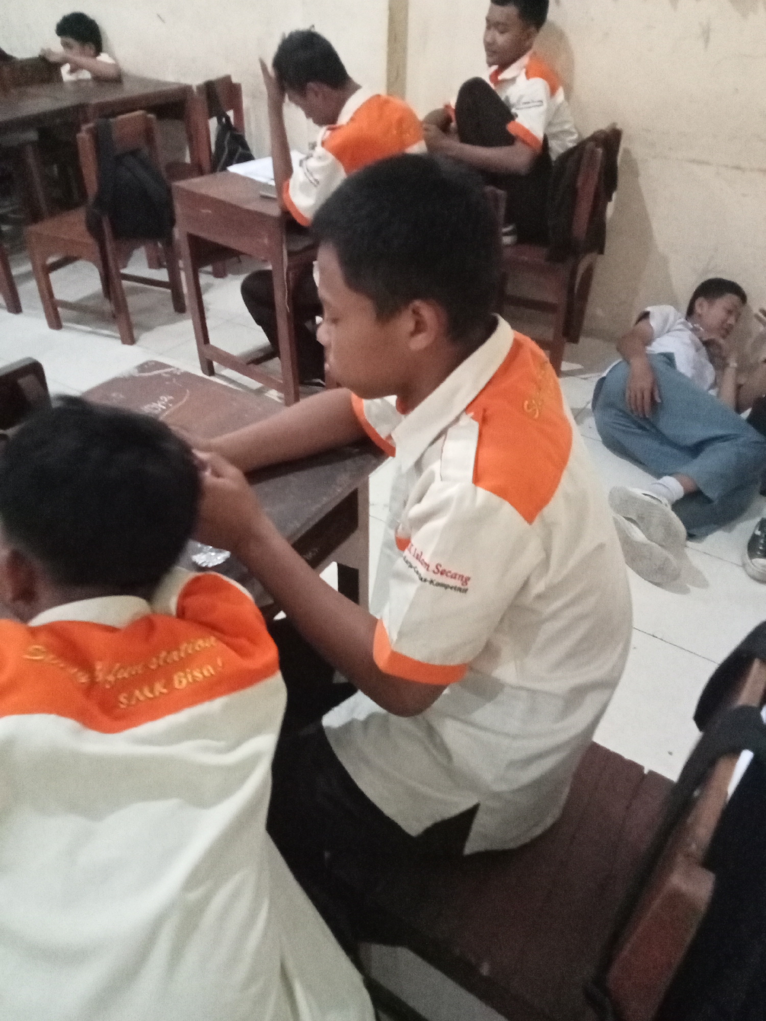 The image depicts a classroom scene at "Sekolah Menengah Kejuruan Islam Secang" (SMK Islam Secang) in Secang, Indonesia. Several young males are present, some seated at wooden desks and chairs, while others are positioned on the floor.
In the foreground, a boy wearing a white shirt with orange accents is visible from the back, facing away from the camera. The shirt has embroidered text, including "Sekolah Islam station" and "SAIK Bisa!".
To the right of the foreground subject, a boy in a similar white and orange shirt is seated at a desk, his head resting on his hand, suggesting fatigue or contemplation. Another boy is seen lying on the floor in the background, wearing a white shirt and blue pants. Further to the left, another young male is seated at a desk, his arm raised.
The environment appears to be a simple classroom with wooden furniture, including desks and chairs. The walls are plain and light-colored. Bags are visible on some of the chairs. The lighting suggests an indoor setting, possibly during the day or with artificial lighting. The overall impression is one of a casual or relaxed school environment.