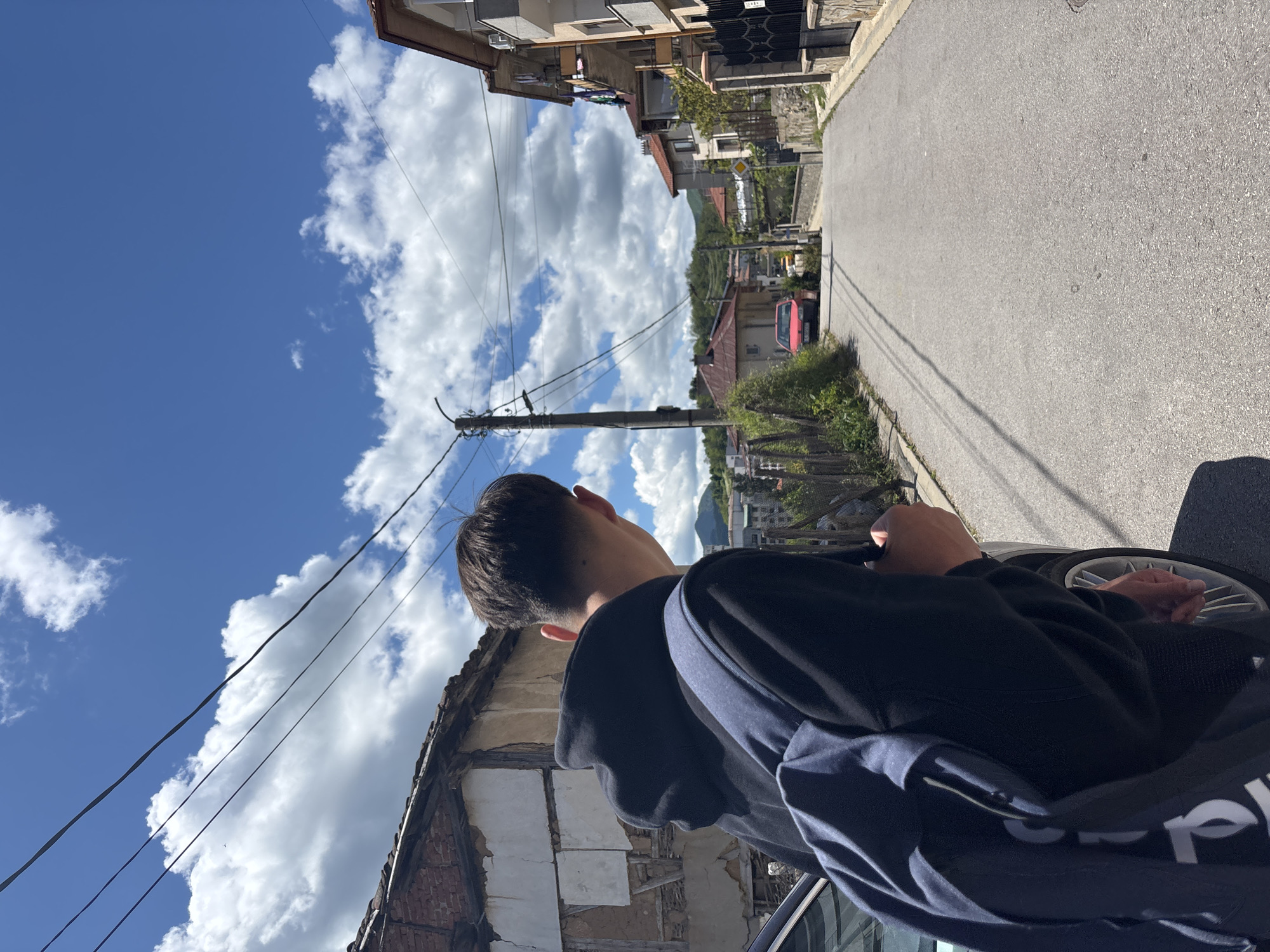 A street view in Zlatograd, Bulgaria, taken in daylight under a sky filled with scattered clouds. A young person with short dark hair and wearing a dark hoodie stands near a car with their hands resting on the steering wheel. The view is oriented towards the sky and the street ahead. A utility pole with wires is visible against the sky, and buildings are seen on both sides of the street. A mountain is visible further down the street. The buildings are of varying heights, and some have visible brick and plaster details. The street is paved and appears slightly downhill.