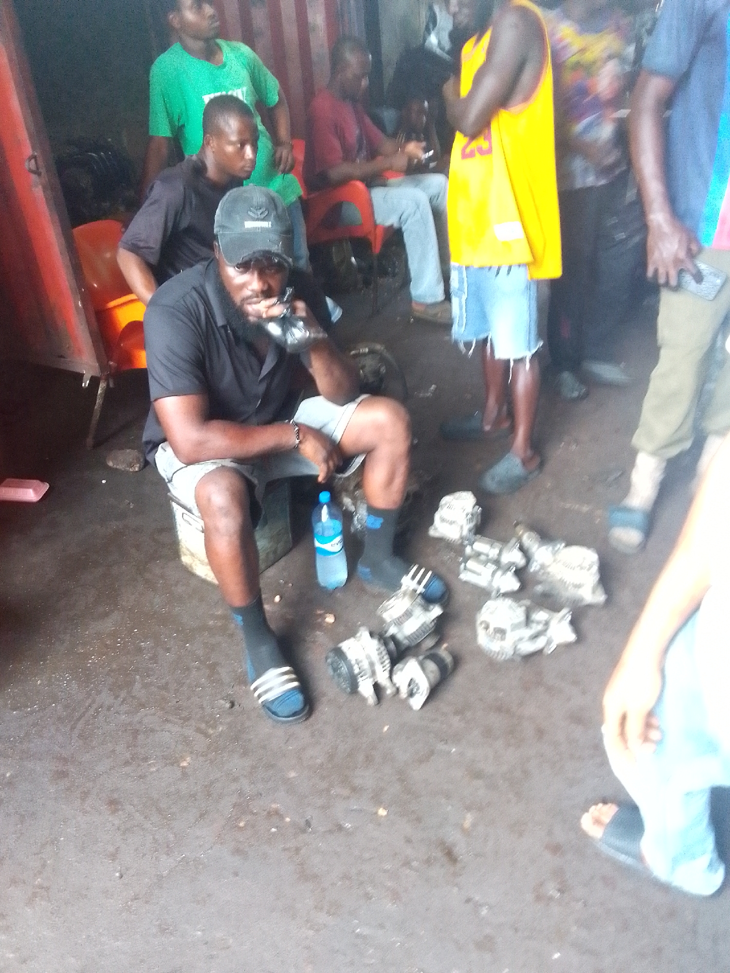 The image captures a scene in what appears to be a mechanic or auto parts area, possibly in Onitsha, Nigeria. A man wearing a black shirt, grey shorts, black socks with blue stripes, and sandals is seated on a low, wooden box. He is holding a black plastic bag up to his face. He has a beard and is wearing a black baseball cap.
Next to him is a half-empty plastic water bottle. In front of him, on the ground, are several car alternators.
Around him stand several other men, some leaning against what appears to be the side of a shipping container. One man is wearing a yellow basketball jersey and denim shorts. Another is in a green t-shirt. Further back, seated on plastic chairs, are other men. The setting is a concrete surface, with evidence of grime and oil. The scene suggests an auto parts market or repair shop.