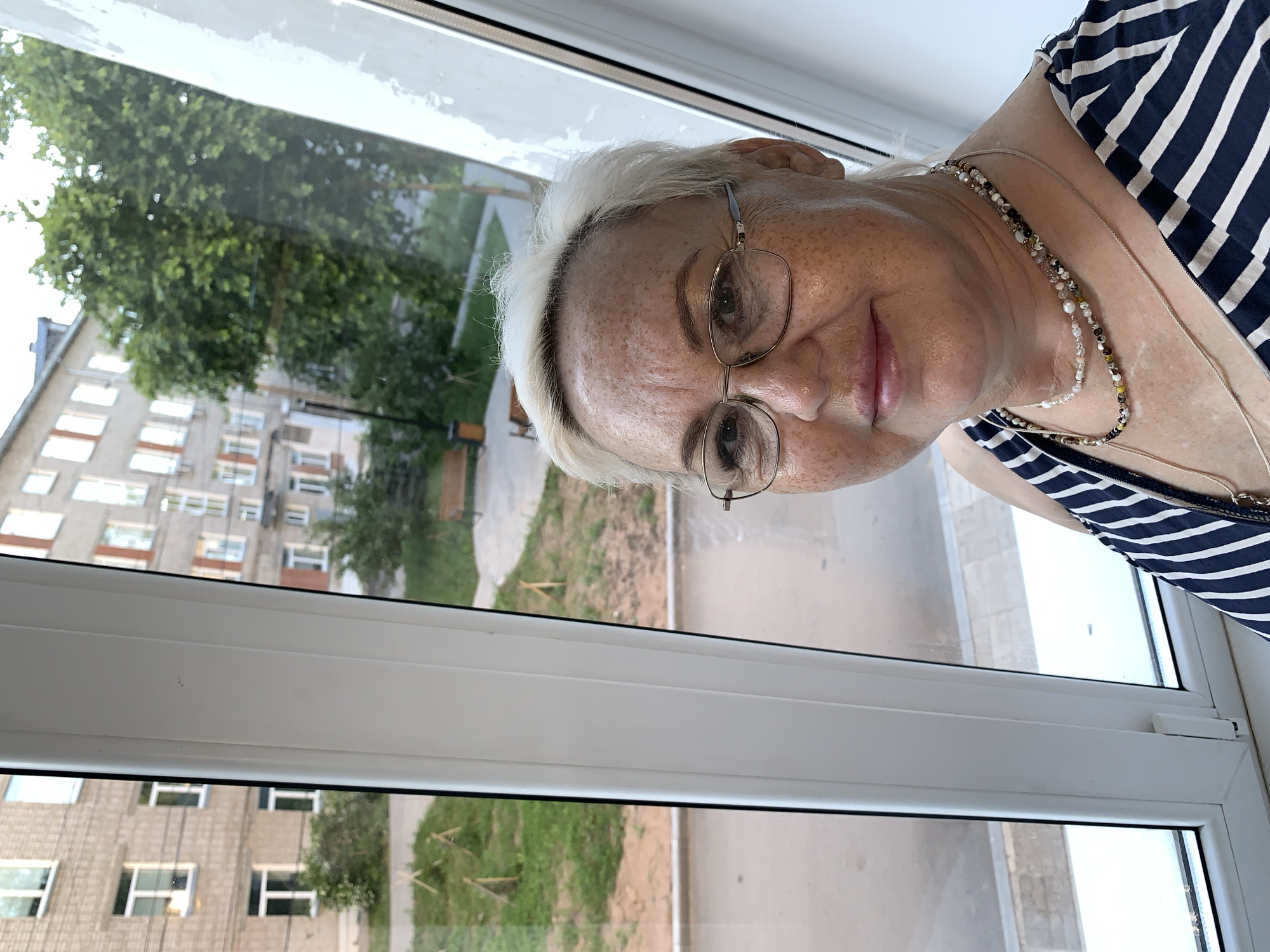 A woman with blonde hair and glasses is taking a selfie in front of a window. She is wearing a striped dress and necklaces. Outside the window, there are apartment buildings and trees, suggesting an urban environment. The scene appears to be a residential area, possibly in Chaikovsky, Russia, given the architectural style of the buildings.