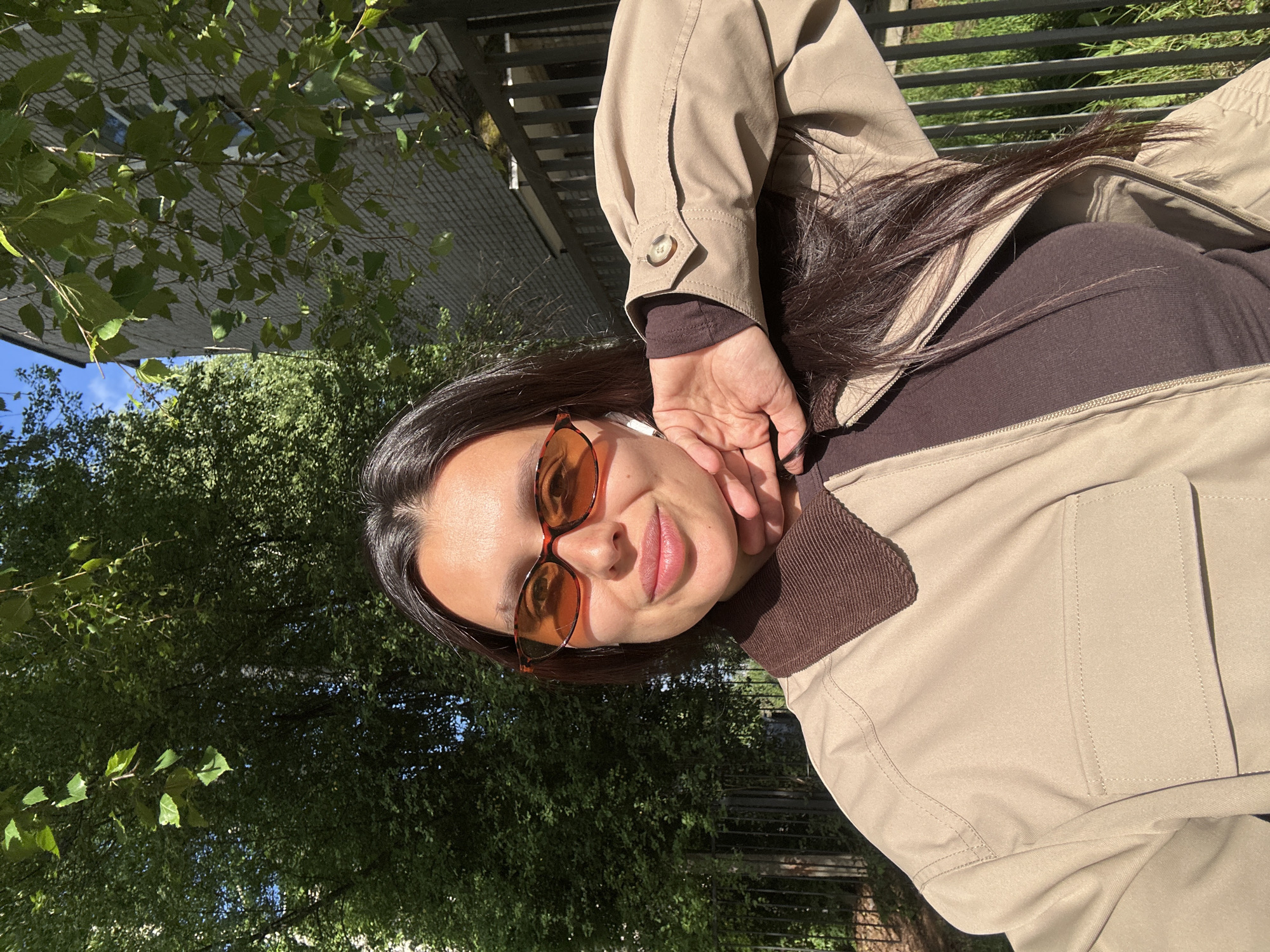The image captures a young woman outdoors in Chaikovsky, Russia, on a bright, sunny day. She is shown from the chest up, positioned at an angle that rotates the view. The woman has dark hair and a fair complexion, and she is looking directly at the camera with a soft, pleasant smile. She wears amber-tinted sunglasses with tortoise-shell frames and has an earbud in her right ear. Her outfit includes a dark brown, possibly ribbed, long-sleeved top and a light beige or khaki jacket with a prominent brown collar. Her right hand is raised, gently resting near her jawline.
The setting is an outdoor environment rich with greenery. Lush green trees and foliage dominate the background, suggesting a park, garden, or a street with abundant vegetation. Behind the natural elements, a grey, structured surface, possibly a building or a sturdy fence, is partially visible. The strong sunlight casts clear shadows, indicating a bright day, likely mid-morning or afternoon. The overall mood is relaxed and natural. There is no visible text in the image.