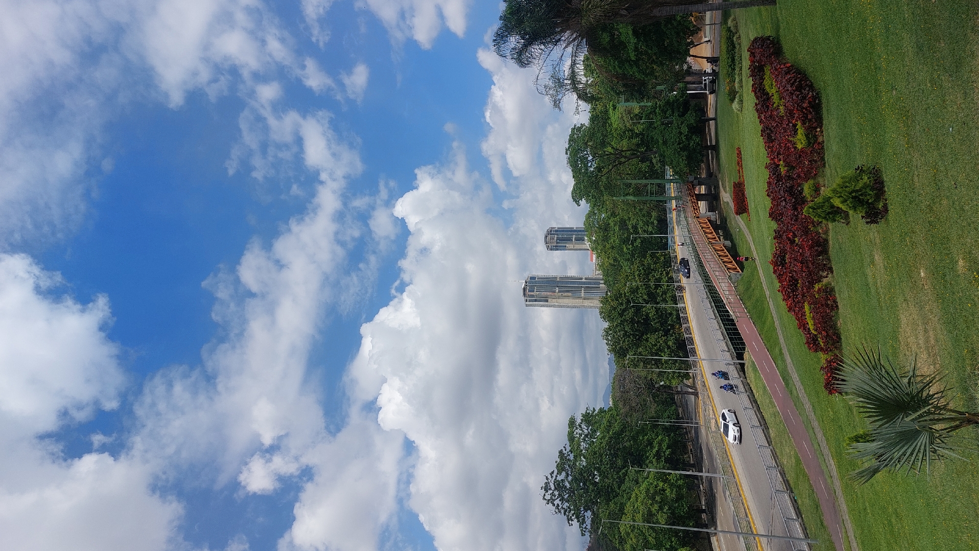 The image shows an elevated view of Caracas, Venezuela. The blue sky is partially obscured by fluffy white clouds. Two tall buildings are visible, their facades reflecting light. A road runs through the scene with cars and motorcycles traveling on it. A pedestrian bridge and a bike path run parallel to the road. Green trees and grassy areas border the road, adorned with red and green ornamental plants.