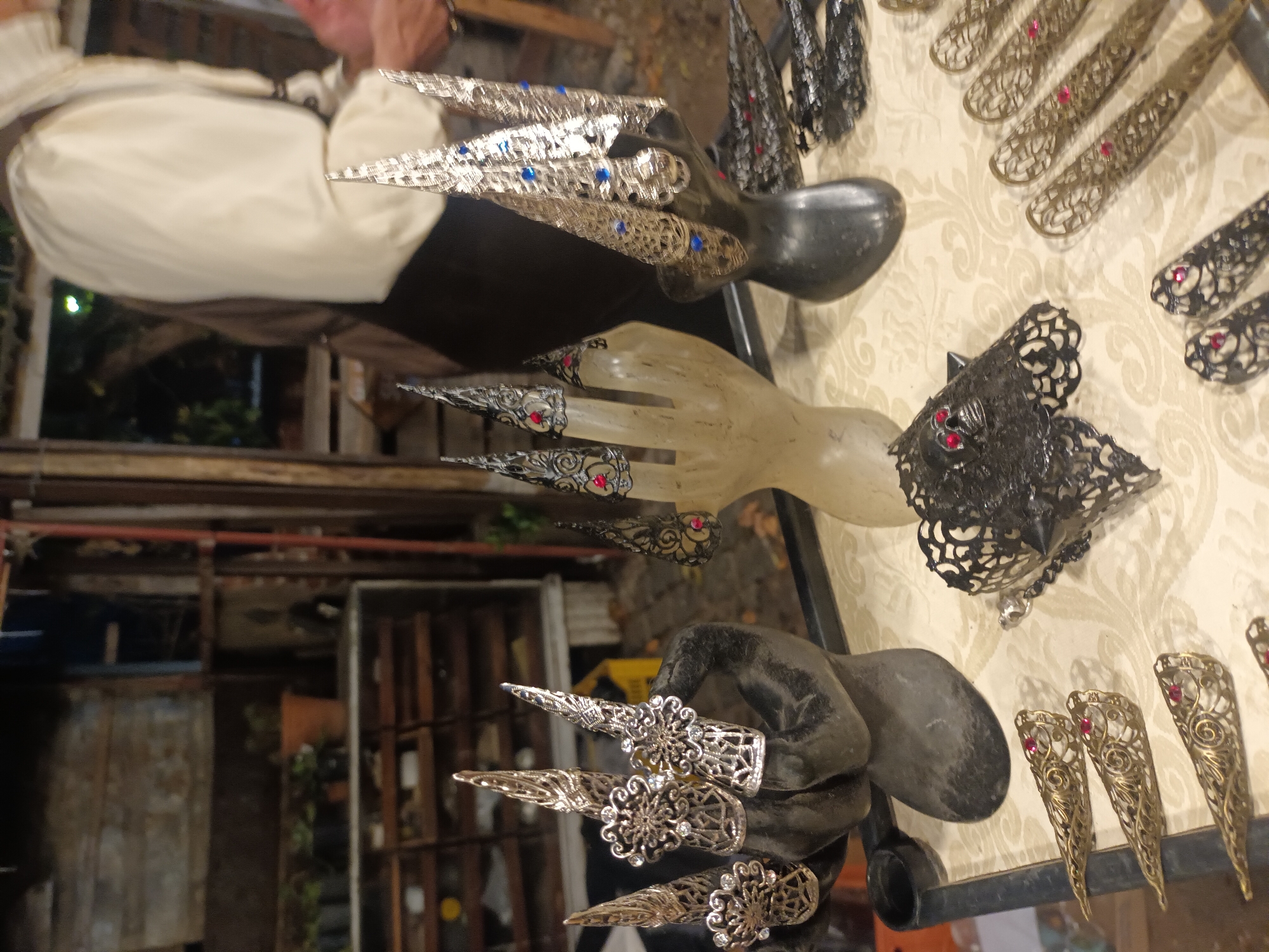 The image displays a collection of decorative finger claws on display at a shop in Buenos Aires, Argentina.
The claws are made of metal and adorned with colored stones, possibly artificial. They are presented on artificial hands for display. The hands are of different colors. One is black, one is white and one is transparent.
A person wearing a white shirt can be seen in the background. The setting appears to be a shop or market. The background is slightly blurred.