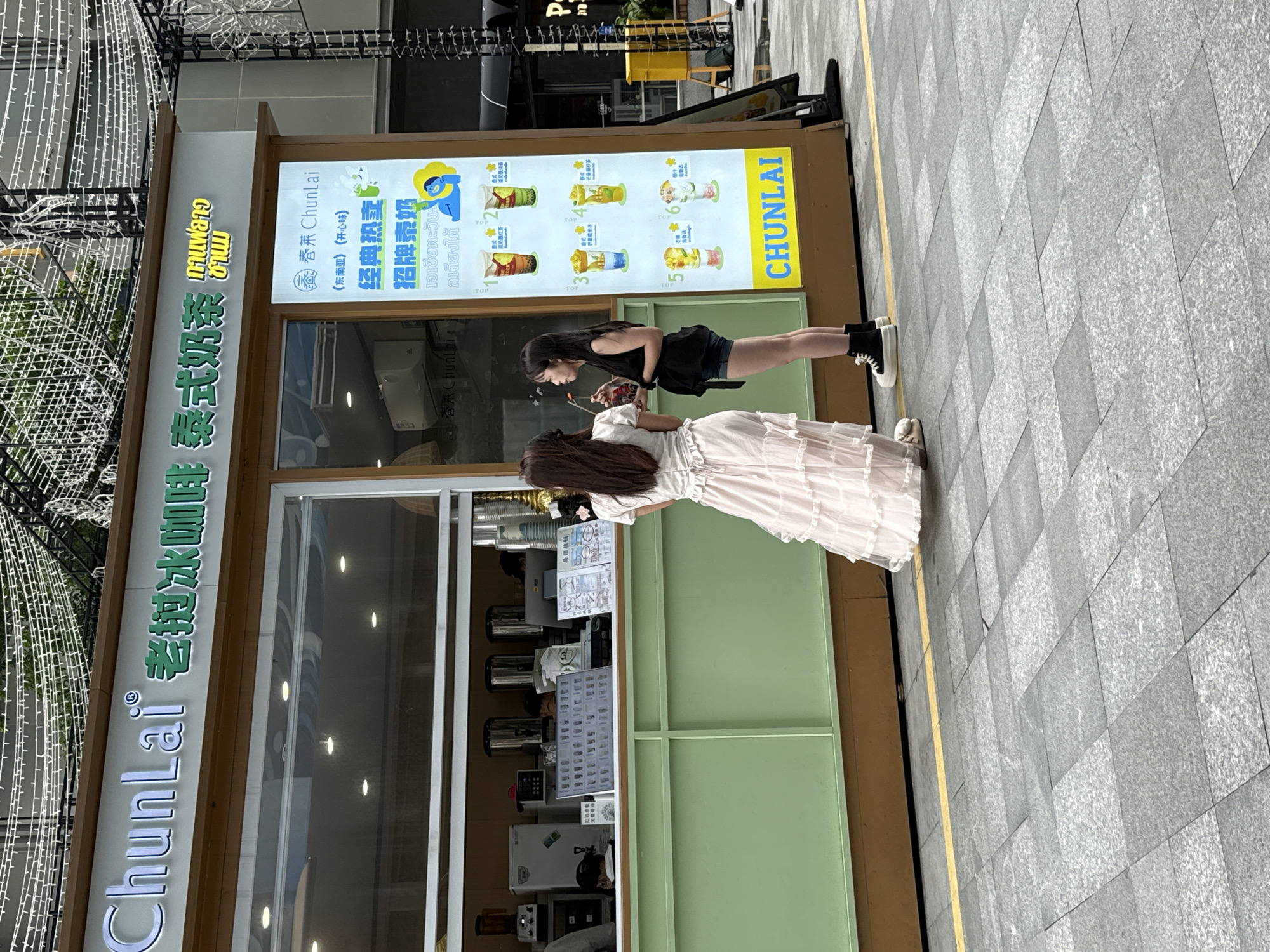 This image captures a street-level view of a "ChunLai" beverage stall, likely located in Huizhou, China, judging by the presence of Chinese characters and the general aesthetic. The stall is an open-air kiosk with a green counter and a wooden trim. Above the counter, a sign displays the brand name "ChunLai" in both English and Chinese characters, along with the words "老挝冰咖啡" (Lao Iced Coffee) and "泰式奶茶" (Thai Milk Tea). To the right of the main sign is a vertical menu board featuring various drinks with Thai text and illustrations of colorful beverages.
In front of the stall, two young women are standing. The woman on the left is wearing a long, flowing pink ruffled skirt and a white top, with her back to the camera. She is holding a drink. The woman on the right is wearing denim shorts and a black top, facing the woman in pink. She is holding a straw and appears to be engaged in conversation with her.
Behind the counter, staff members can be seen working, with equipment such as blenders, kettles, and a refrigerator visible. The ground in front of the stall is paved with grey granite tiles, and a yellow line marks a boundary. The background shows a street scene with more buildings and decorative string lights overhead, suggesting a commercial or pedestrian area in the city.