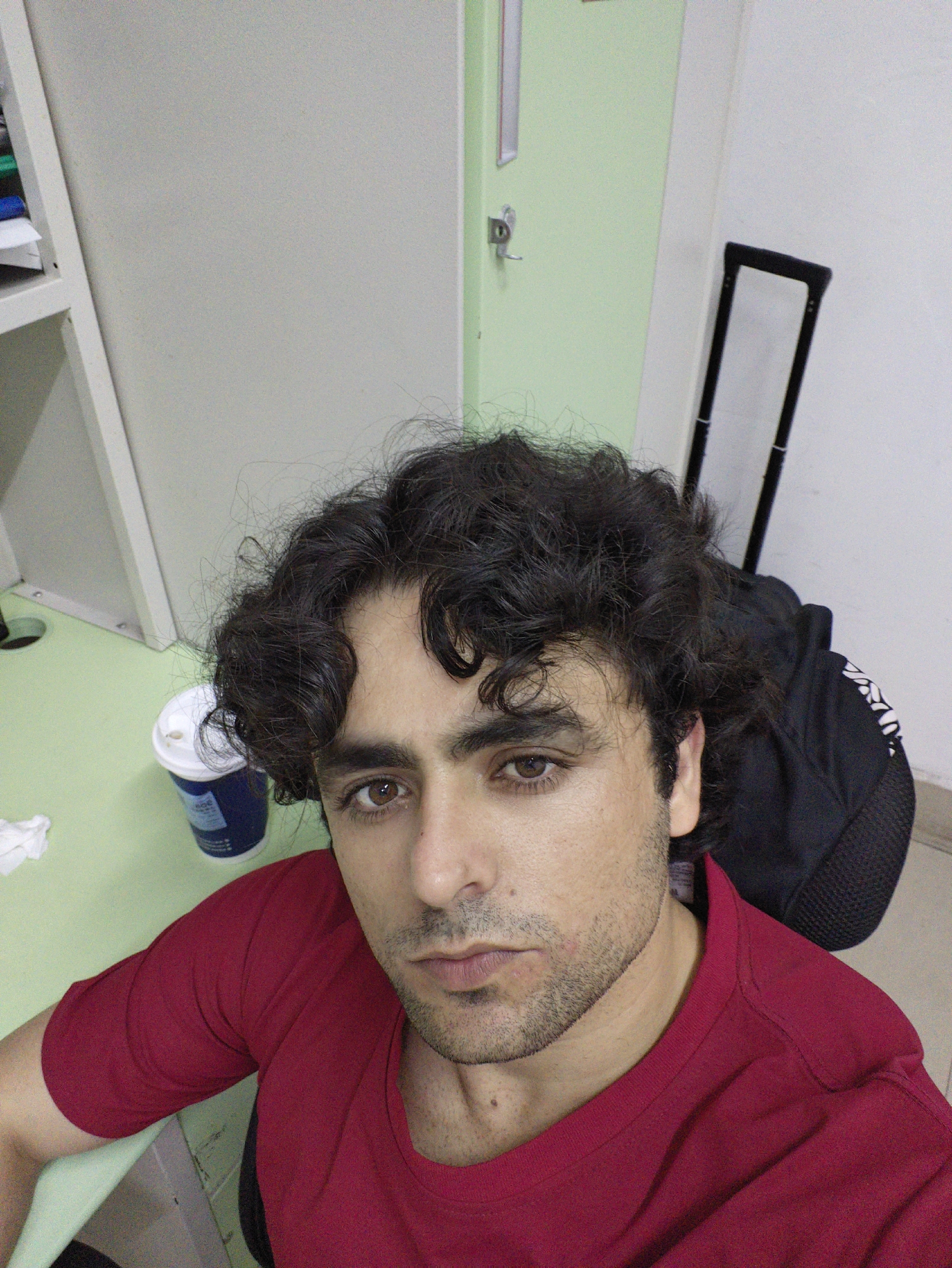 A close-up, high-angle selfie of a man with curly dark hair and brown eyes, wearing a red t-shirt, is taken in what appears to be an office or study environment. The man's face is the central focus, with his head slightly tilted. To his left, a light green desk is visible, with a blue and white disposable cup and a crumpled white tissue. Behind him, a tall, light green door with a silver handle and lock is slightly ajar, revealing a white wall and a black rolling suitcase with a silver handle. The overall impression is casual, possibly taken during a break or at home. The question asks for details about the location of Chongqing High-tech Zone, China, however, there is no visible information or context within the image to support this claim.