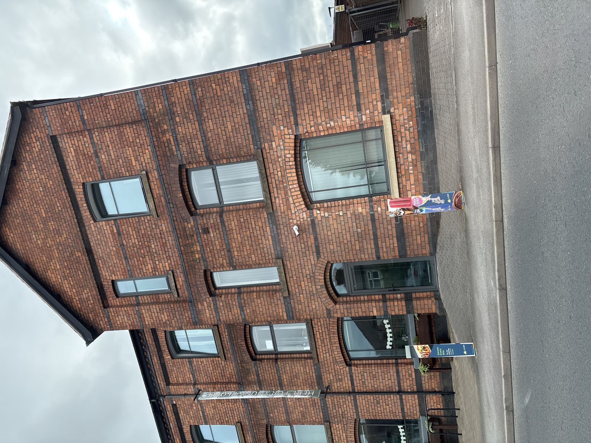 A three-story brick building is depicted in this image, located in Crewe, United Kingdom. The building has a dark, sloped roof and numerous windows with dark frames. A vertical banner with white text reading "HERE FOR YOUNG PEOPLE" is attached to the left side of the building. Two signs are placed on the sidewalk in front of the building; one is blue and advertises food with the text "Savour Every Bite" and "Try our Nation's Chicken." The other sign appears to be a colorful advertisement for food items like ice cream. The building is situated next to a paved walkway and a street. The sky is overcast with clouds. There are no people visible in the image.