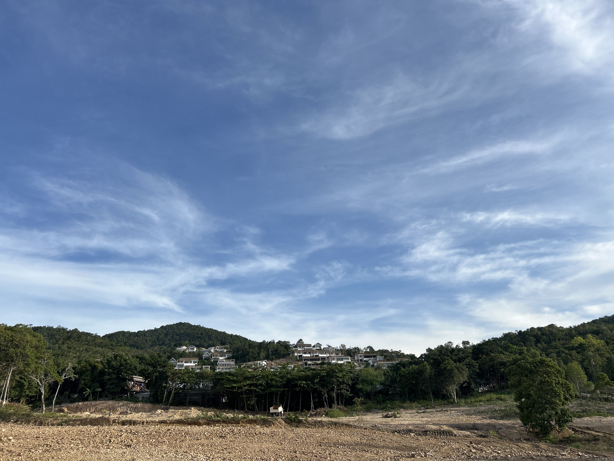 The image shows a landscape in Baan Wang Ta Kien, Thailand. In the foreground, there is a dry, rocky area. A line of trees separates the foreground from the hillside. Several houses are built on the hillside. Behind the hillside, there is a partially cloudy blue sky.