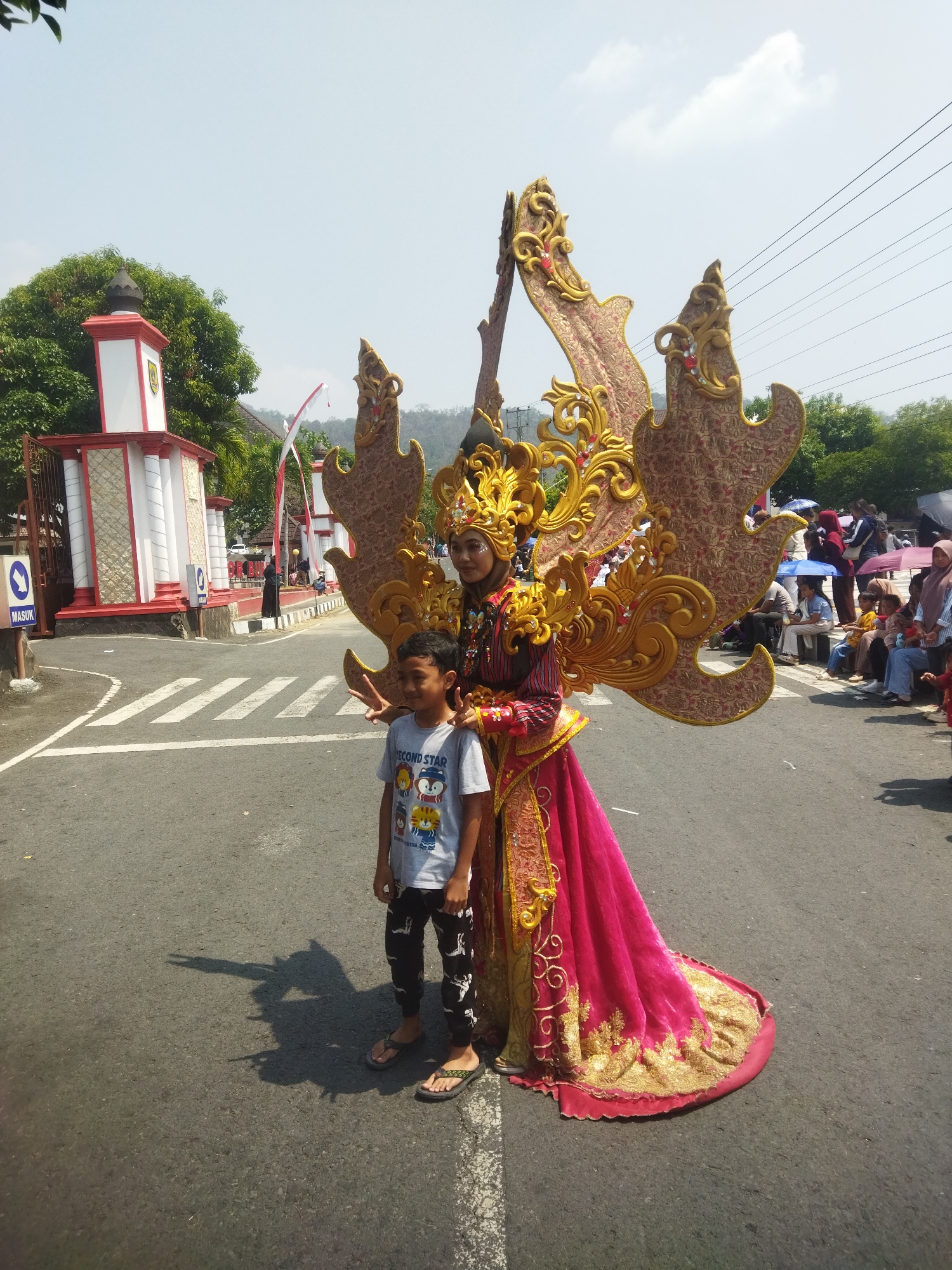 In Wonogiri, Indonesia, on a sunny day, a boy stands next to a woman in an elaborate, golden, and red costume, likely participating in a cultural parade. The woman wears a headdress and a large, winged ornament on her back, suggesting a performance or celebratory event. The background shows a street with a pedestrian crossing, a building with red and white architectural elements, trees, and a crowd of spectators sitting on the side of the road, some holding umbrellas. Power lines are visible against the clear blue sky. The atmosphere appears festive, with onlookers gathered to witness the procession.