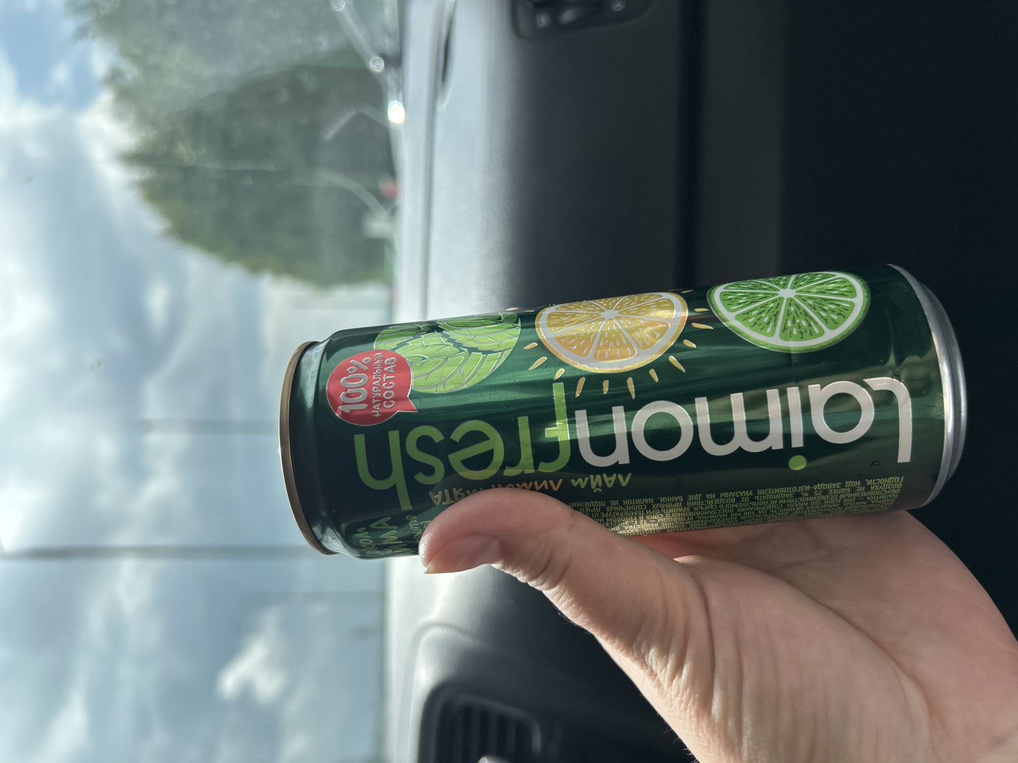 In Arkhangelskoye, Russia, a hand holds a green aluminum can of "Laimon Fresh" beverage. The can is adorned with images of lime and lemon slices, along with mint leaves. A red circular label on the can reads "100% НАТУРАЛЬНЫЙ СОСТАВ" (100% Natural Ingredients). The background suggests the can is inside a vehicle, with the cloudy sky and trees visible through the windshield. The interior of the car, including the dashboard and controls, is also partially visible.