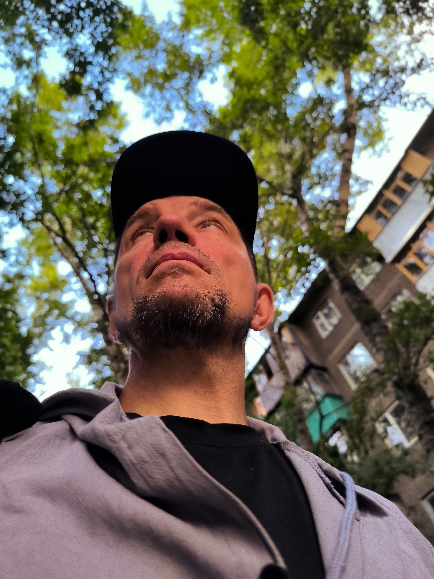 A low-angle, close-up shot focuses on the upper body of a man wearing a black baseball cap and a light purple hooded jacket over a black t-shirt. He is looking upwards, with his face illuminated by sunlight. The background is blurred, showing green foliage from trees against a bright sky, and a portion of a multi-story building with windows. The image appears to be taken outdoors in an urban setting, possibly in Qarağandı city administration, Kazakhstan, given the architecture. The man's skin tone suggests he may be Caucasian. He has short, dark hair, stubble on his chin, and a prominent nose and lips. His left ear is partially visible. The lighting creates a dramatic effect, with shadows and highlights on his face and clothing. The overall impression is one of casual observation or contemplation of the sky and surroundings.