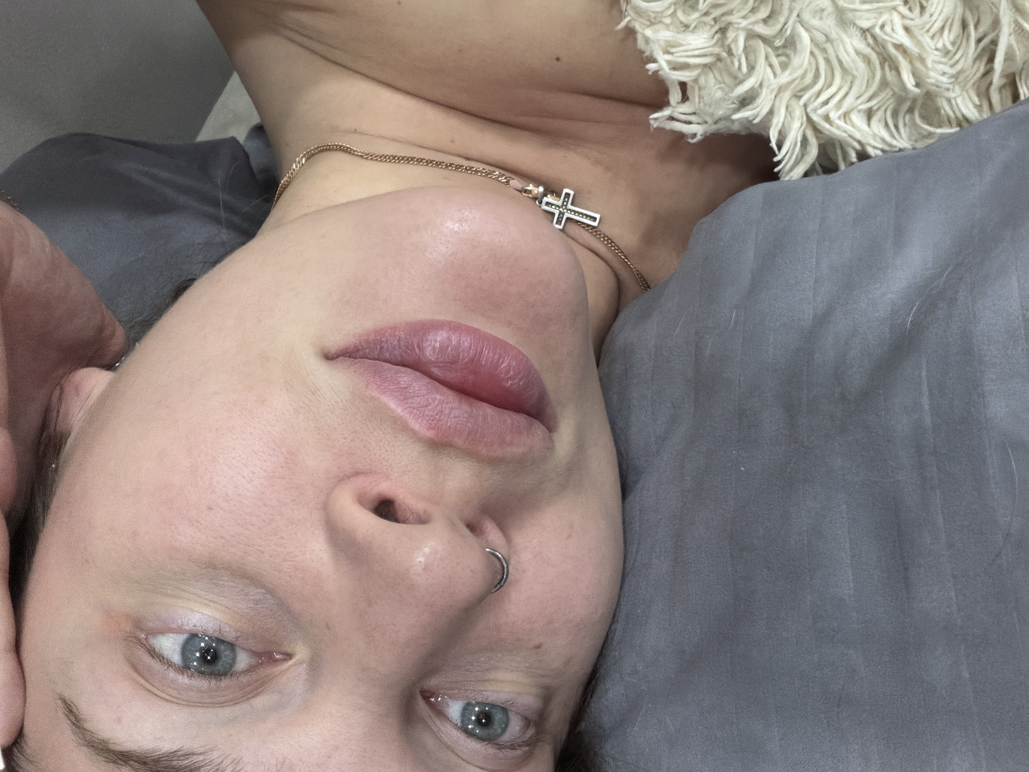 A selfie shows a person lying on gray bedding with a white furry textile visible. The person has light skin, blue eyes, a nose ring, and is wearing a gold necklace with a cross pendant. The lips are slightly parted. The photograph does not offer any information about the city of Chaikovsky, Russia.