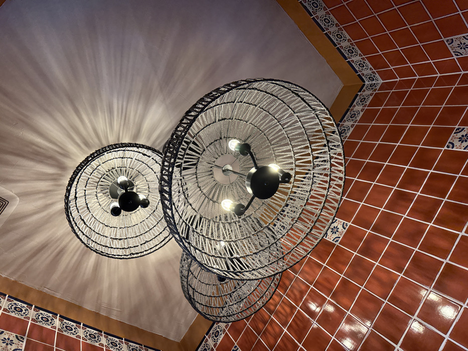 The image depicts an upward view within the interior of Miguel's Cocina in Chula Vista, United States of America. Three large, circular ceiling light fixtures dominate the frame. Each fixture consists of a dark metal framework with multiple exposed incandescent bulbs, surrounded by a wide, woven shade crafted from light-colored cord or yarn wrapped around a dark circular rim. The uppermost fixture casts distinct radial light patterns onto the pale, textured ceiling. The upper portions of two interior walls are visible, meeting at a corner. These walls are clad in uniform reddish-brown square tiles with light-colored grout lines. A decorative border, featuring small patterned blue and white tiles topped by a solid ochre-yellow band, runs along the junction of the tiled walls and the light-colored ceiling. No individuals, actions, or interactions are present in the scene.