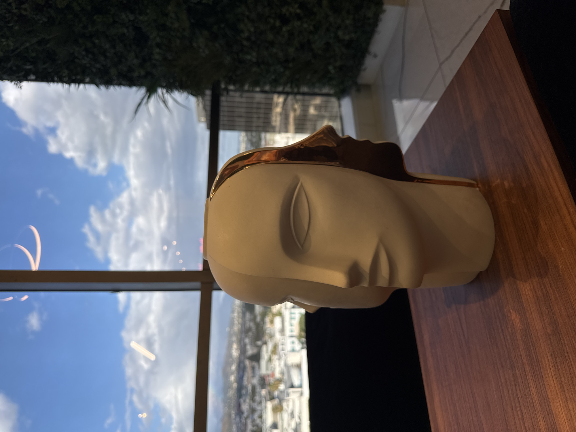 The image features a bust sculpture of a face, white with gold accents, positioned on a wooden surface. Outside a window, a view of a city skyline can be seen, along with a clear blue sky with clouds. The presence of lush greenery is visible at the top of the frame. Based on the architecture and landscaping visible through the window, it can be inferred that the location is Beverly Hills, United States of America.