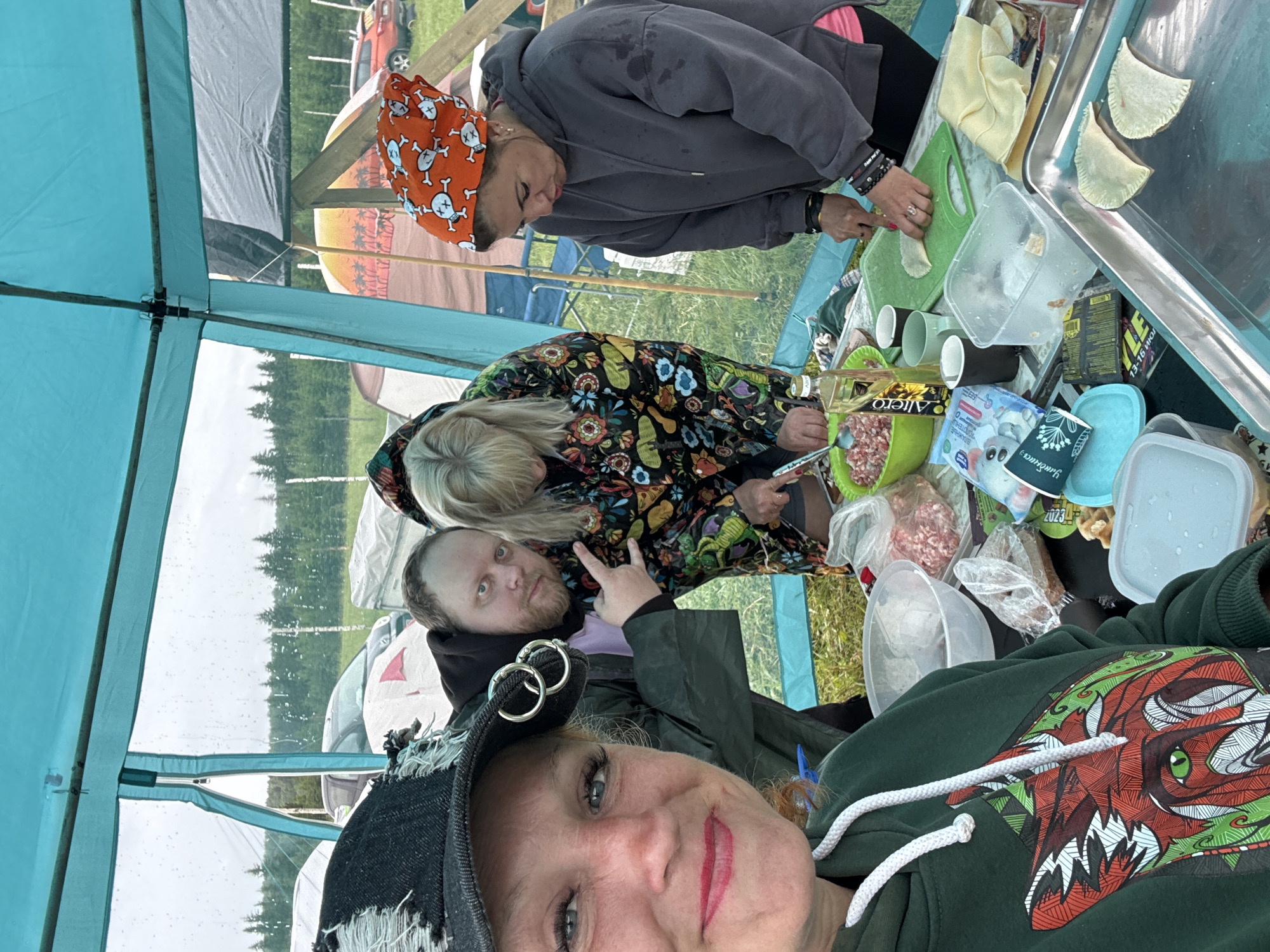 A group of four people is engaged in outdoor food preparation under a temporary canopy or tent structure, likely at a campsite in Большая Кивара, Russia. The image is a selfie taken by a woman in the foreground, who looks directly at the camera with red lipstick and a slight smile. She wears a dark green hooded sweatshirt featuring a prominent red, white, and green stylized animal or monster head graphic, and a dark baseball cap with two silver rings.
Behind her, a man with a beard makes a peace sign towards the camera. To his right, a woman with long blonde hair is focused on preparing food, wearing a colorful patterned jacket with floral and possibly insect designs. Further back, another woman, wearing a grey hooded sweatshirt with visible pink lining and an orange bandana adorned with white skull and crossbones patterns, is actively working on a green cutting board. She is seen pressing or shaping dough, with several semi-circular pastry items already arranged on a metal tray next to her.
The makeshift cooking area includes various items: bowls of ground meat, a bottle of "Allegro" cooking oil, plastic containers, disposable cups (some white, some black), a package labeled "Универсальная" (Universal), and another package with "2023" visible. A small box also shows "13-16 ИЮЛ" (July 13-16).
The setting appears natural, with a line of pine trees visible in the background through the clear panels of the tent. Water droplets on these panels suggest recent rain or current light drizzle. Other tents, camping chairs, and a red vehicle are faintly visible in the distance. The atmosphere seems relaxed and cooperative, suggesting a shared activity during an outdoor gathering or camping trip.
