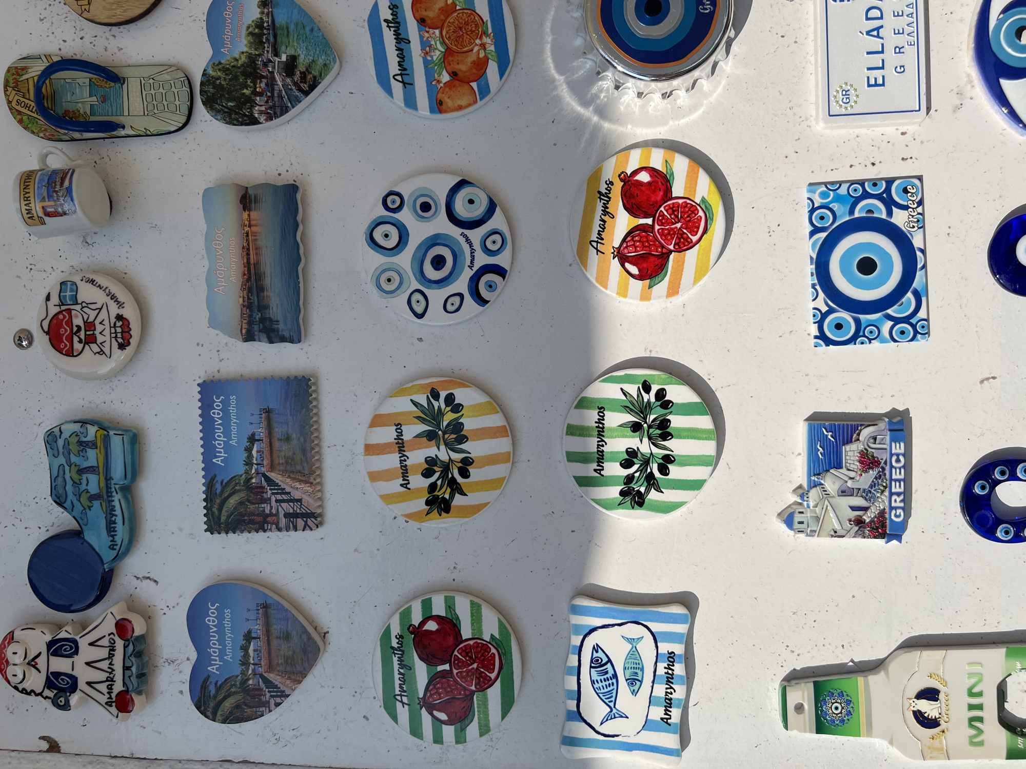 The image is a close-up, top-down view of a collection of assorted souvenir magnets adhered to a white, textured surface, likely a wall or refrigerator. The magnets depict various motifs, including local imagery, traditional patterns, and whimsical designs.
In the upper left quadrant, a magnet shaped like a stylized figure wearing a blue and white outfit with red accents is present, with "AMARYNTHOS" written below. Adjacent to it is a magnet shaped like a blue shoe with a depiction of palm trees and a body of water, also labeled "AMARYNTHOS." To the right of this, a round magnet features a cartoon character with a red hat and a Greek flag. Next to this is a small white mug-shaped magnet with a lighthouse scene and "AMARYNTHOS" on it. Further right is a flip-flop shaped magnet with a sailboat and a coastal scene.
In the middle row, a heart-shaped magnet displays a scenic view of a waterfront with palm trees and the text "Αμάρυνθος" and "Amarynthos." To its right is a rectangular magnet resembling a postage stamp, also featuring a waterfront scene and "Αμάρυνθος" and "Amarynthos." Below this, a rectangular magnet shows a coastal town at sunset with "Αμάρυνθος" and "Amarynthos." To the far right, another heart-shaped magnet has a similar waterfront scene.
The bottom two rows contain more diverse magnets. There are several circular magnets with striped backgrounds and illustrations of pomegranates, olive branches, and oranges, all labeled "Amarynthos." Another magnet is shaped like a plaque with blue and white stripes and illustrations of two fish, labeled "Amarynthos." A magnet shaped like a bottle with a cat and "Greece" is present in the lower left. To its right is a magnet depicting a classic Greek island village with white buildings and blue domes, labeled "GREECE." In the lower middle is a rectangular magnet covered in a repeating pattern of blue and white evil eye symbols, with "Greece" written on it. To the right of this is a white rectangular license plate-style magnet with "ELLÁDA" and "GREECE" printed on it. Several other round magnets with evil eye designs are also visible.
The lighting suggests it is daytime, with some shadows casting across the surface. The overall impression is a collection of tourist souvenirs, predominantly related to Amarynthos and Greece.