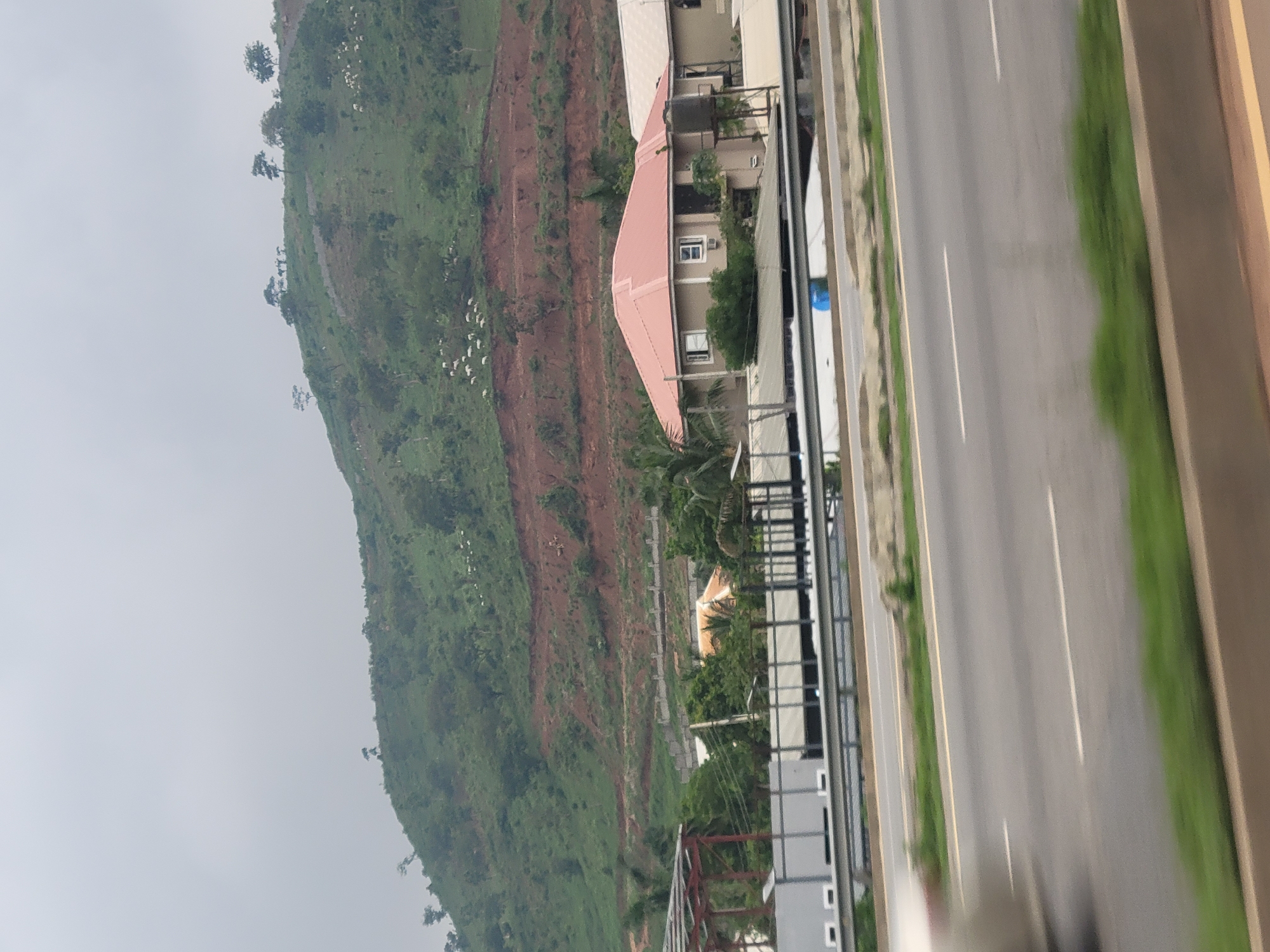 In Kugbo, Nigeria, a hillside with sparse vegetation and a large area of exposed reddish-brown soil is visible under a hazy sky. A herd of white animals, likely sheep, are scattered across the green slopes. Several trees are present on the hillside. In the foreground, a multi-lane highway runs horizontally across the image, with a concrete barrier and green verges. Behind the highway, a beige house with a salmon-colored roof stands out. Several other structures, including what appears to be an unfinished building with metal framework and some walls, are visible behind the house. The scene suggests a developing area with a mix of natural landscape and construction.