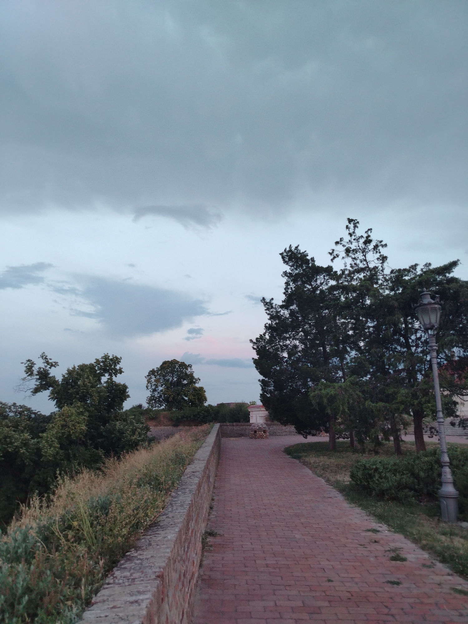 The image depicts a pathway made of red brick, curving to the right, leading into the distance. The pathway is lined on the left by a low brick wall, beyond which is overgrown vegetation and trees. On the right side of the pathway, there are more trees and a vintage-style lamppost. The sky above is overcast with clouds. The scene appears to be an outdoor public space, possibly a park or a promenade, with a sense of tranquil, natural scenery. The location is identified as the Petrovaradin Fortress in Petrovaradin, Serbia.