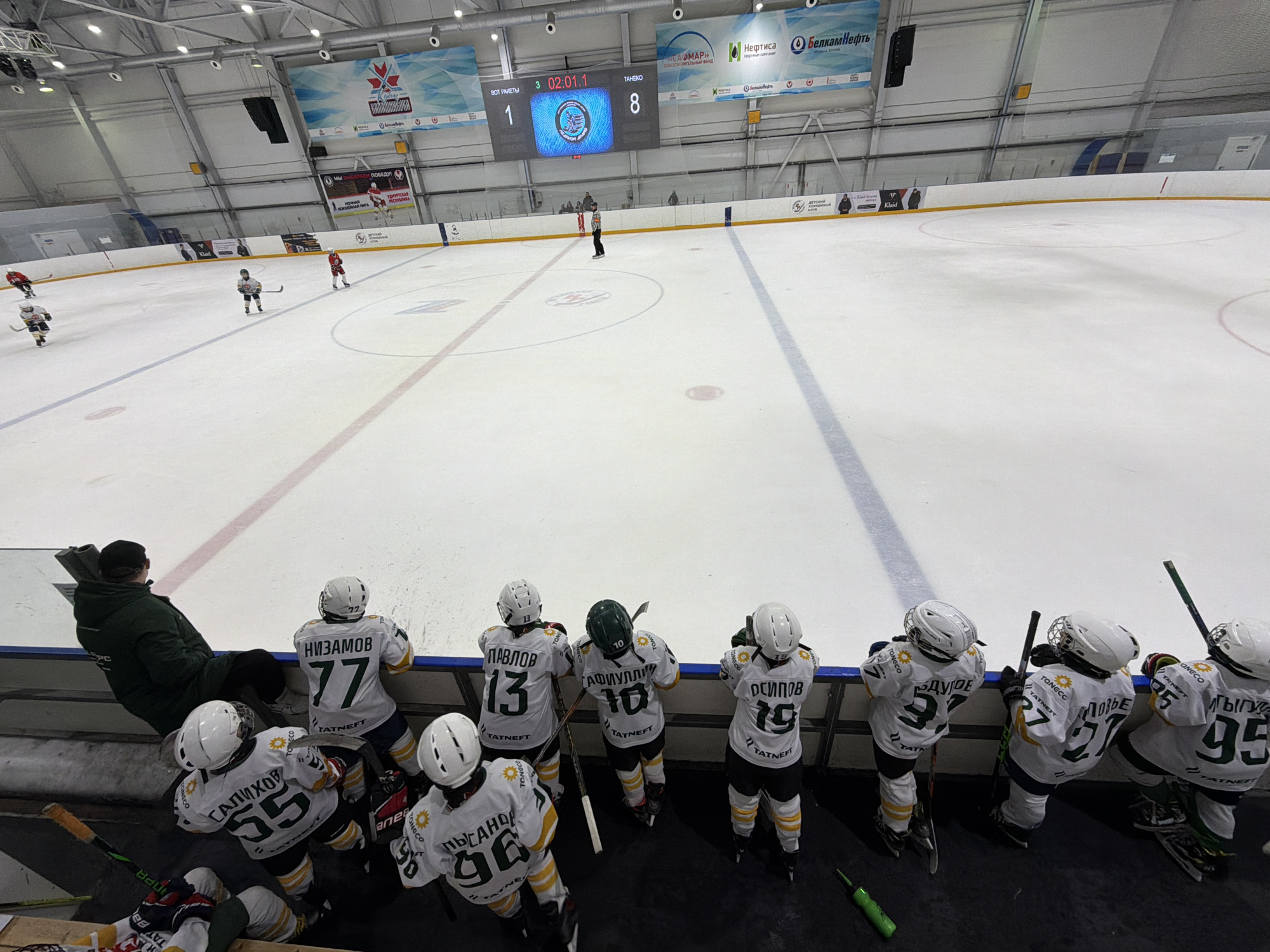 Here is a description of the image:
The image shows a hockey game in progress at an indoor arena, likely in Votkinsk, Russia. The scoreboard indicates the game is in the third period with the score being 1-8. Several young hockey players in white and green jerseys are sitting on the bench, observing the game. The players have numbers such as 77, 13, 10, 19, 37, 55, 96, and 95 on their jerseys, along with the logo of "TATNEFT". A coach is seated at the end of the bench, also watching the game. The arena is well-lit, and there are advertising banners visible in the background. There are players visible on the ice, actively participating in the game.