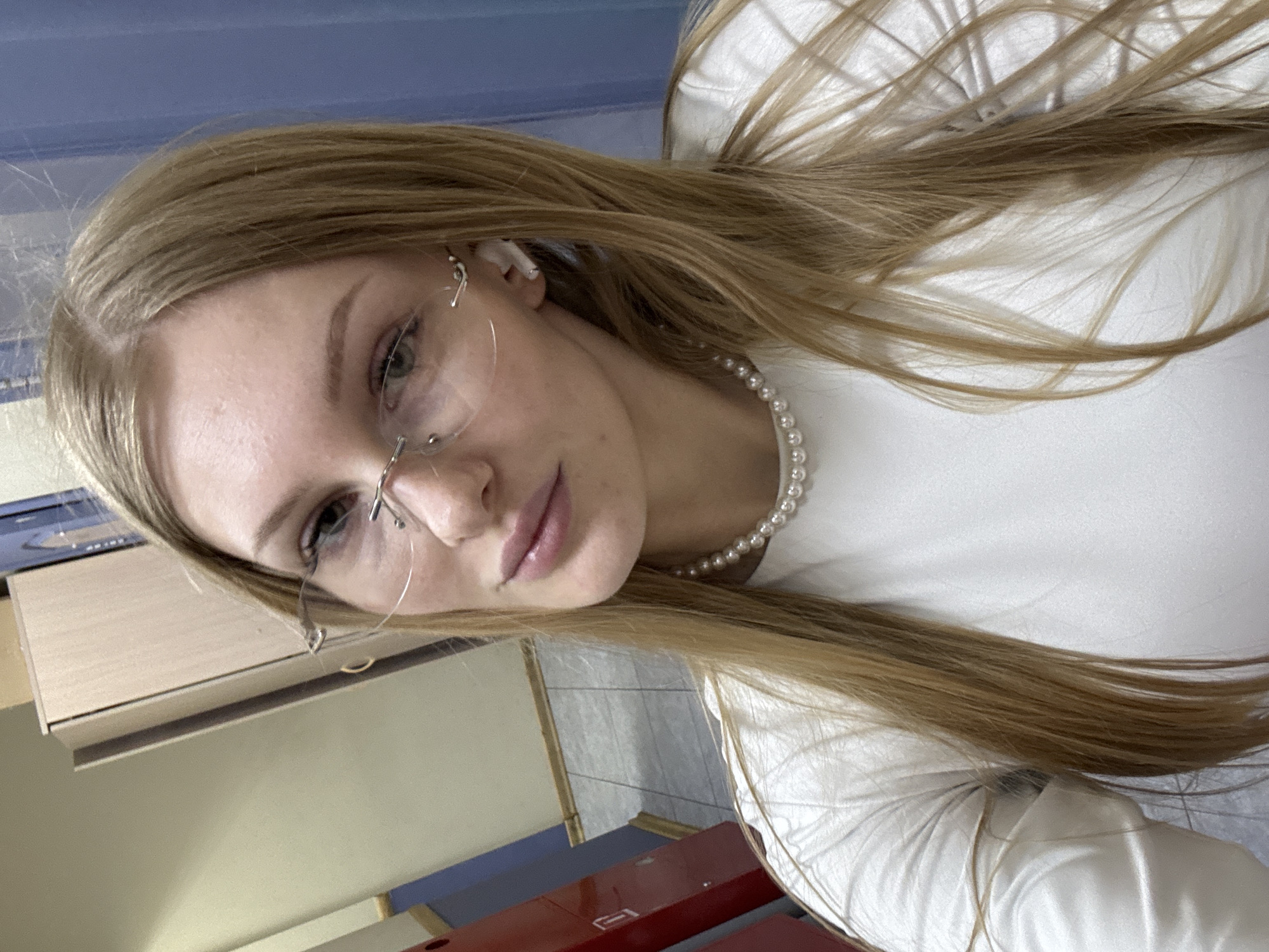 A close-up, tilted portrait features a young woman with long, straight, light brown hair. She is wearing a simple white top and a pearl necklace. Rimless eyeglasses are perched on her face, and a white AirPod is visible in her right ear. Her lips are slightly parted, and she has a neutral to serene expression while looking towards the camera. The background suggests an indoor setting, with blurred elements including light-colored wooden furniture, blue and grey wall sections, and what appears to be a tiled floor. A glimpse of a red object is visible in the lower right. The lighting indicates it is daytime. There is no visible text in the image. While the location context is Perm, Russia, this is not discernible from the visual cues within the photograph.