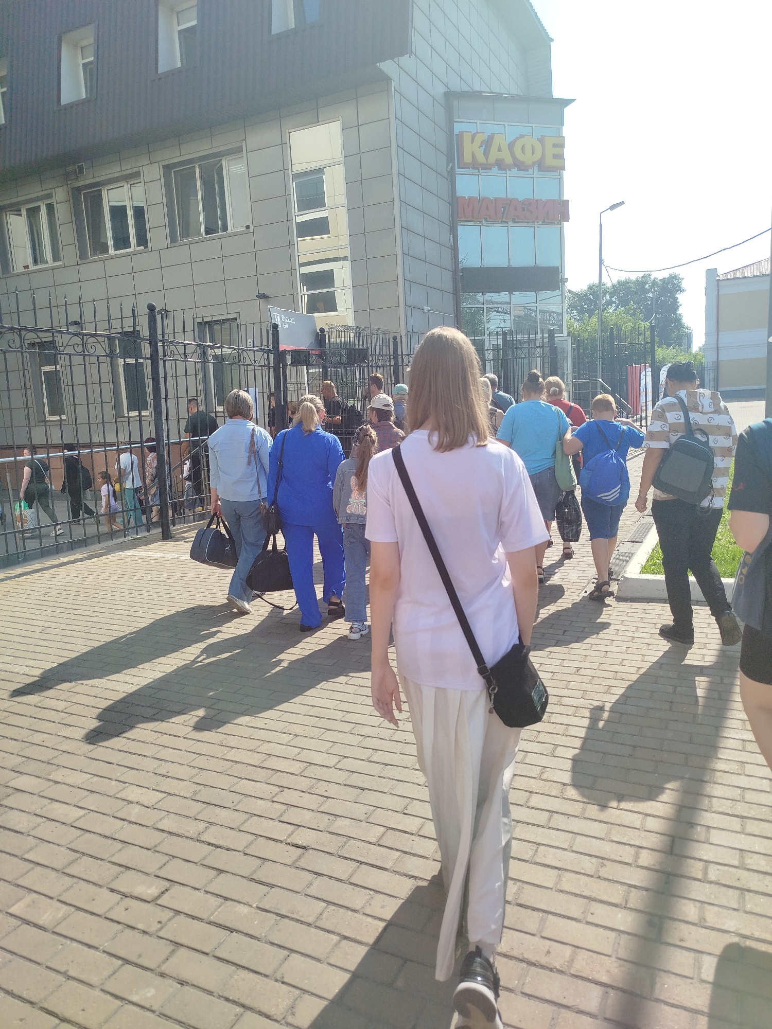 In the city of Tyumen, Russia, a crowd of people walks on a brick-paved sidewalk in front of a building with a sign that reads "KAFE MAGIZIN" (Cafe Store). The building has a modern facade with large windows. Several individuals are carrying bags, suggesting they may be shoppers or travelers. The sun is casting long shadows on the ground, indicating it is likely daytime. The atmosphere appears casual, with people moving in the same direction.