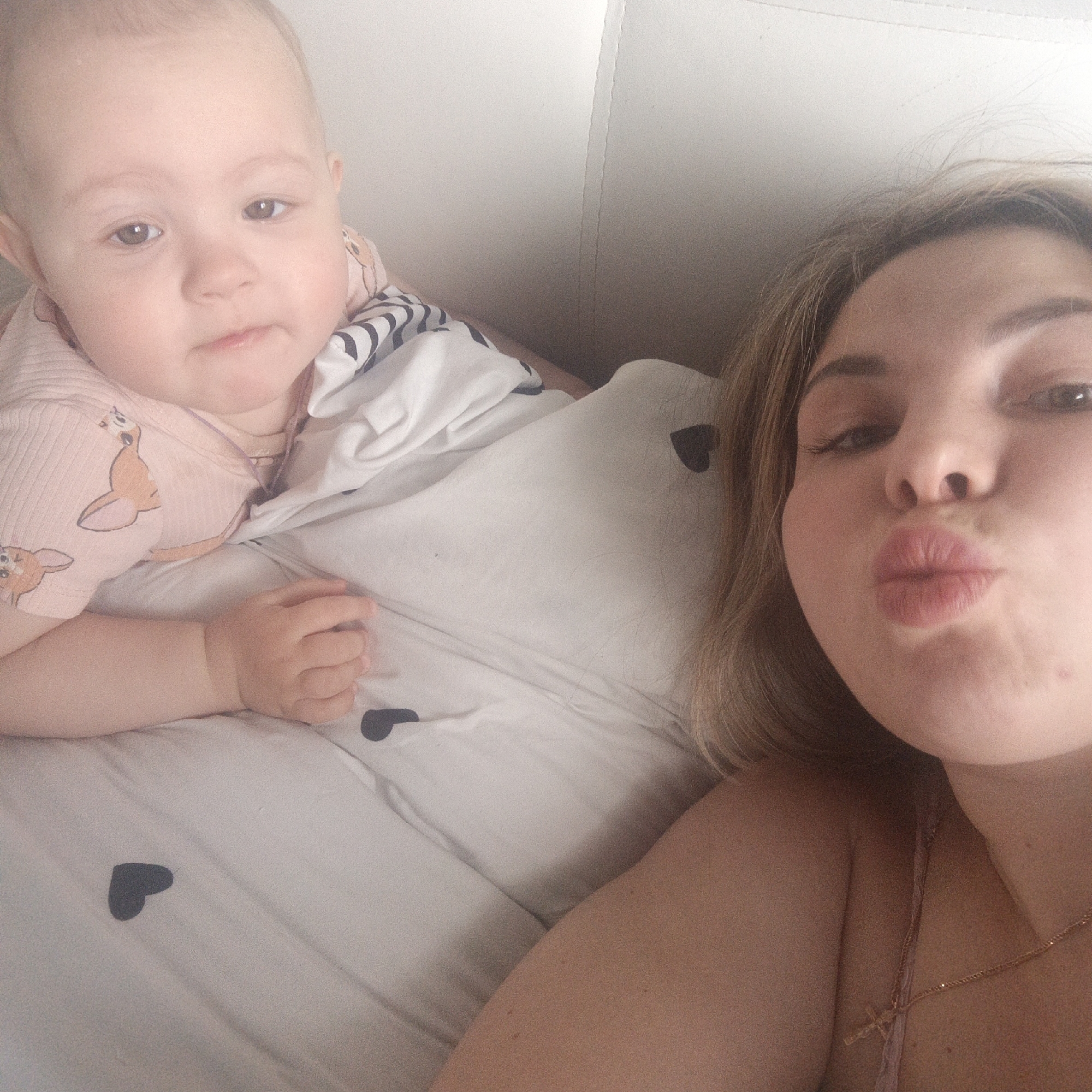 A selfie shows a woman with blond hair pursing her lips, lying in bed next to a baby. The baby is lying on its stomach, wearing a pink shirt with a Bambi pattern. The bed has a white comforter with black hearts on it. The location is Unknown, Unknown.