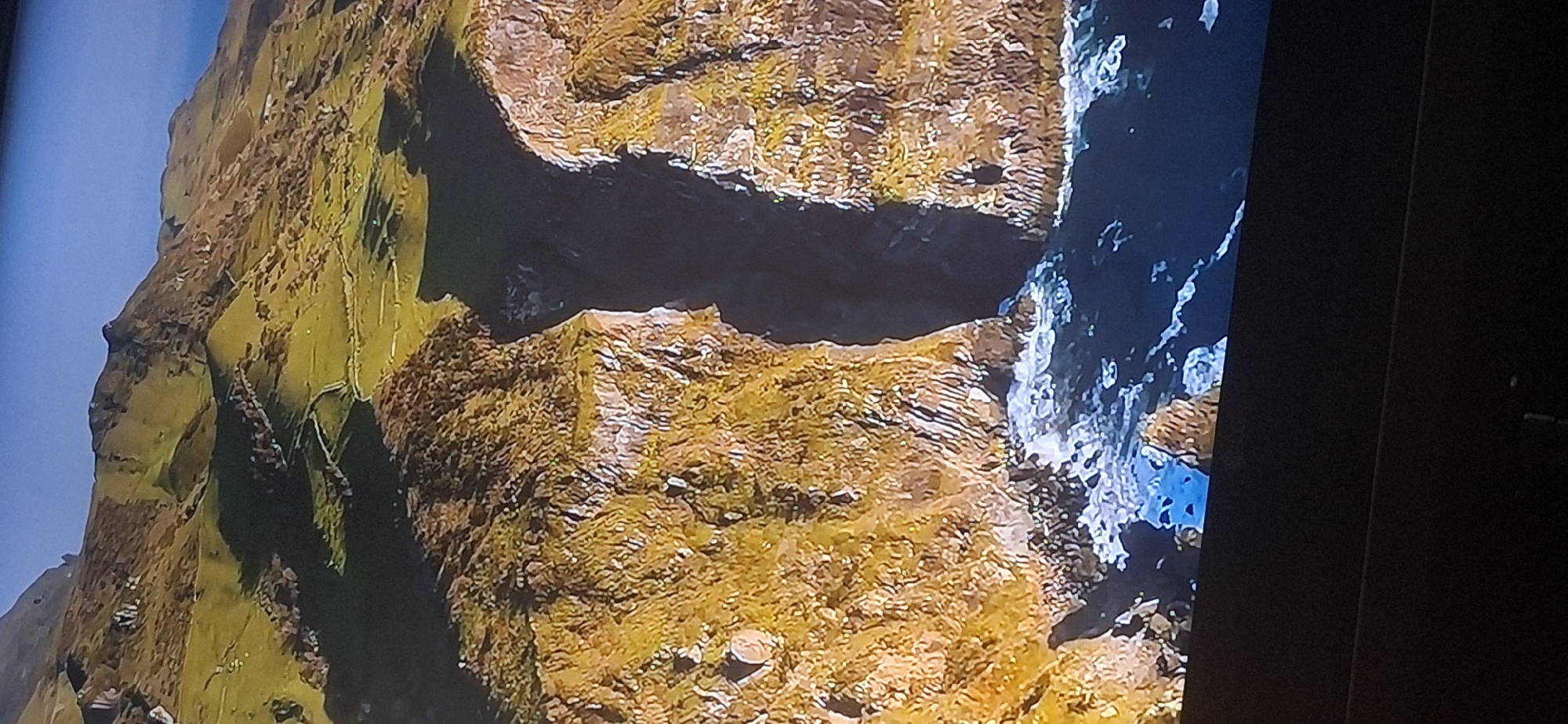 The image shows a drone's perspective of a rugged coastal landscape with steep cliffs, grassy hills, and the ocean. The cliffs are dry and rocky, covered with patches of yellow and green vegetation, indicative of a Mediterranean or similar climate. The hills above are a vibrant green, with winding paths or roads visible, suggesting human activity and potentially agricultural use. The ocean below is dark blue with white waves crashing against the base of the cliffs, creating a sense of natural power and erosion. The lighting suggests it is daytime, possibly late afternoon due to the long shadows cast on the hills. The image does not depict any urban environment or specifically the city of Bila Tserkva, Ukraine; rather, it is a natural landscape.