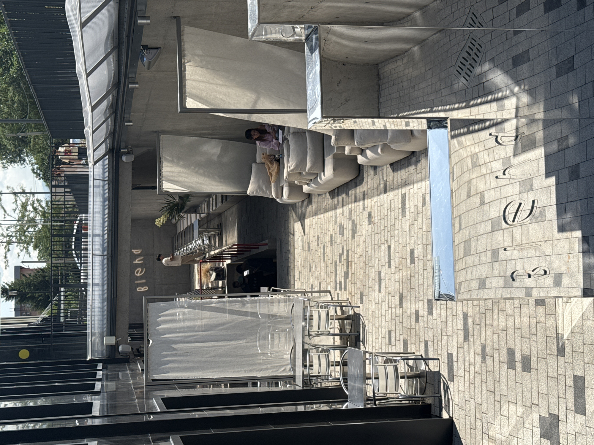 In Almaty, Kazakhstan, an outdoor seating area features a modern design with concrete walls and a translucent roof. Several tables with metal chairs are set up on a brick-patterned ground. On the right side, a person sits on a large, plush sofa, reading a book. The scene is bathed in sunlight, casting shadows across the area, with a building facade on the left and trees visible in the background. The word "blend" is visible in white lettering on the concrete wall.