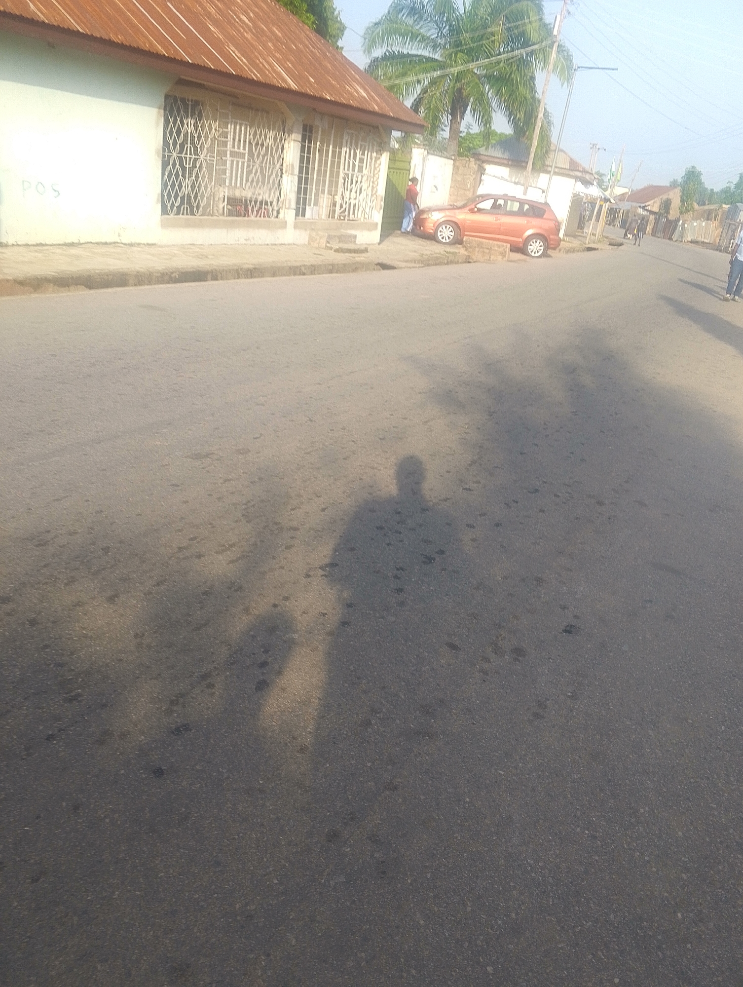 The image depicts a street scene in Jalingo, Nigeria. In the foreground, the asphalt road is visible, with the shadow of the photographer and possibly another person cast upon it. The shadow of a person's head and shoulders is prominently displayed on the left side of the frame. In the background, a light-colored building with a corrugated metal roof and barred windows is situated alongside the road. A light orange hatchback car is parked on the side of the road in front of this building. To the right of the building, a palm tree stands, and further down the street, there are other structures and people walking. The sky is bright, indicating daylight. The overall impression is of a typical urban street environment.