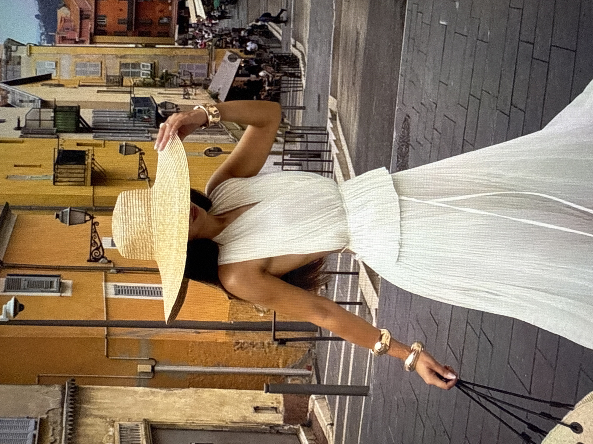 A woman wearing a white, sleeveless, V-neck dress with a peplum waist and a pleated skirt is walking down a cobblestone street. She has on a large straw hat and gold bracelets on her wrists. She is holding a black-strapped bag in her left hand. The background features buildings with yellow and orange facades, balconies, and windows. The street is lined with people, suggesting a lively urban environment. The scene is reminiscent of a Mediterranean town, but the prompt specifies the location as Găvănoasa, Moldova.