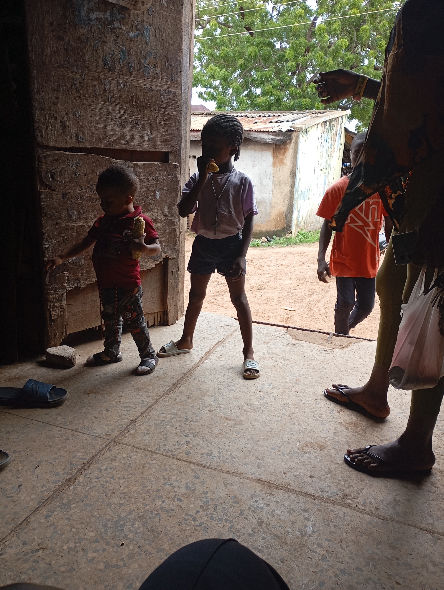 The image depicts a scene within a dwelling or structure in Shendam, Nigeria. The foreground shows a person's legs and feet wearing black flip-flops, with another person's legs and feet also visible nearby, one wearing brown flip-flops. A white plastic bag hangs from the right side of the frame.
In the middle ground, two young children stand on a concrete floor. The child on the left, a boy wearing a red shirt and patterned pants, holds a piece of corn. He is looking down and to his left. The child on the right, a girl wearing a light purple t-shirt and dark shorts, is eating a piece of corn. Her hair is styled in braids.
Behind the children, a young person wearing an orange t-shirt and dark pants walks away from the camera. The background shows a rustic building with a corrugated metal roof, and trees with green foliage, suggesting an outdoor or semi-outdoor environment. The overall setting appears to be a rural or semi-urban area, consistent with the location of Shendam, Nigeria. The structure in the foreground is made of weathered wood, with visible grain and some discoloration. The ground is paved with speckled concrete tiles.