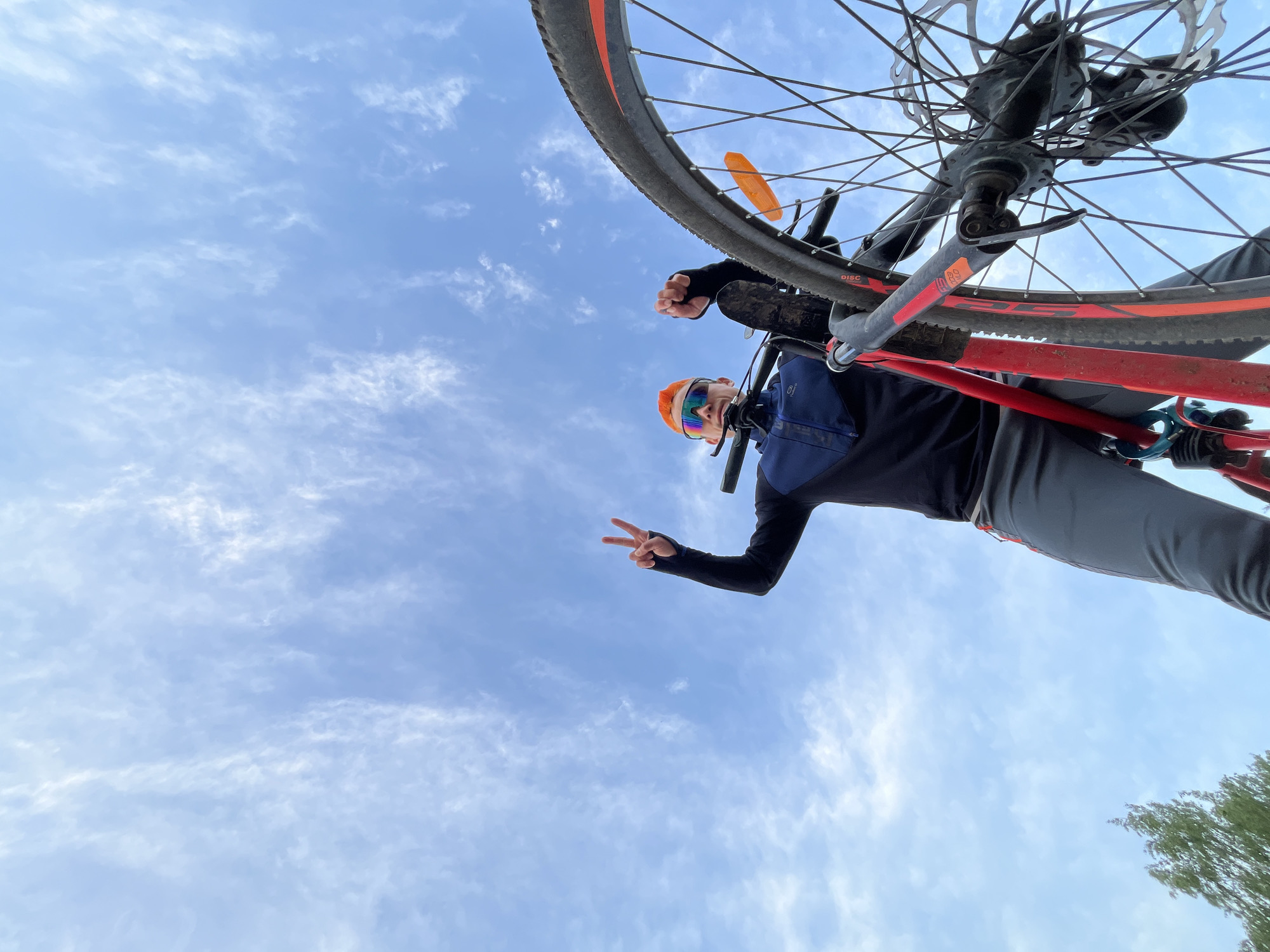 A low-angle shot captures a cyclist standing with their bicycle, holding it above them against a backdrop of a partly cloudy sky. The cyclist, wearing a black long-sleeved shirt, grey pants, an orange helmet, and sunglasses, is holding up two fingers in a peace sign. The bicycle is positioned above the cyclist, with the front wheel and fork dominating the upper part of the frame. The bicycle's frame is predominantly red. In the bottom right corner, tree branches are visible against the sky. The location is the city of Chaikovsky, Russia.