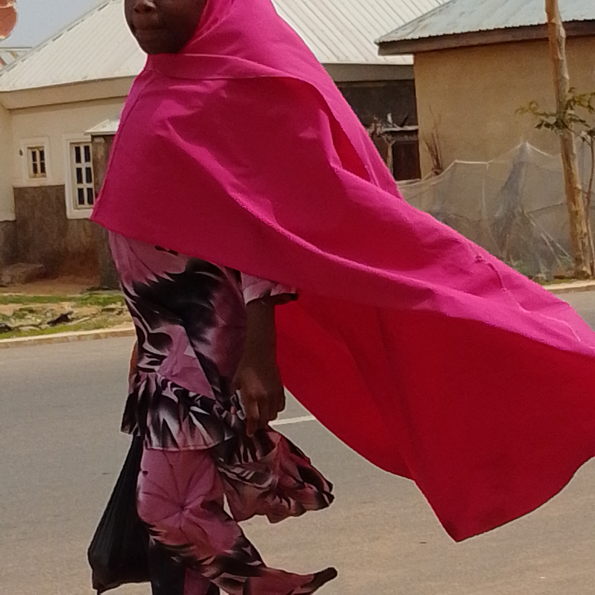In Gombe, Nigeria, a person wearing a bright pink hijab and a patterned pink and black dress is captured mid-stride on a paved road. The fabric of the hijab billows dramatically, creating a dynamic visual element. The dress features a floral or abstract pattern in shades of pink and black, with a ruffled hemline. The person's face is partially visible, and their dark skin tone is apparent. The background shows a low-rise building with white trim and windows, and a tall, thin tree to the right. The environment suggests an outdoor, possibly residential, setting within the city of Gombe. The lighting indicates a sunny day.