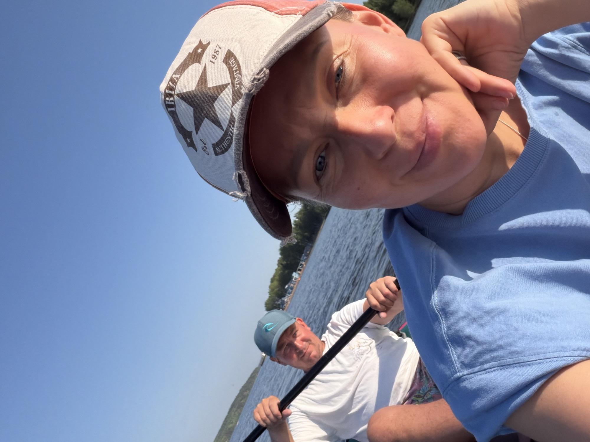 A daytime outdoor shot shows two people on the water. The primary subject, closer to the camera, is a light-skinned person wearing a light blue top and a tan baseball cap with a star and the word "VINTAGE" on it. They are looking at the camera with a slight smile. Behind them, a second person, also light-skinned, is holding a paddle and wearing a white shirt and a blue baseball cap. They are also looking towards the camera. The background features a body of water and a distant shoreline with trees. It is impossible to ascertain whether this image was taken in the city of Chaikovsky, Russia.